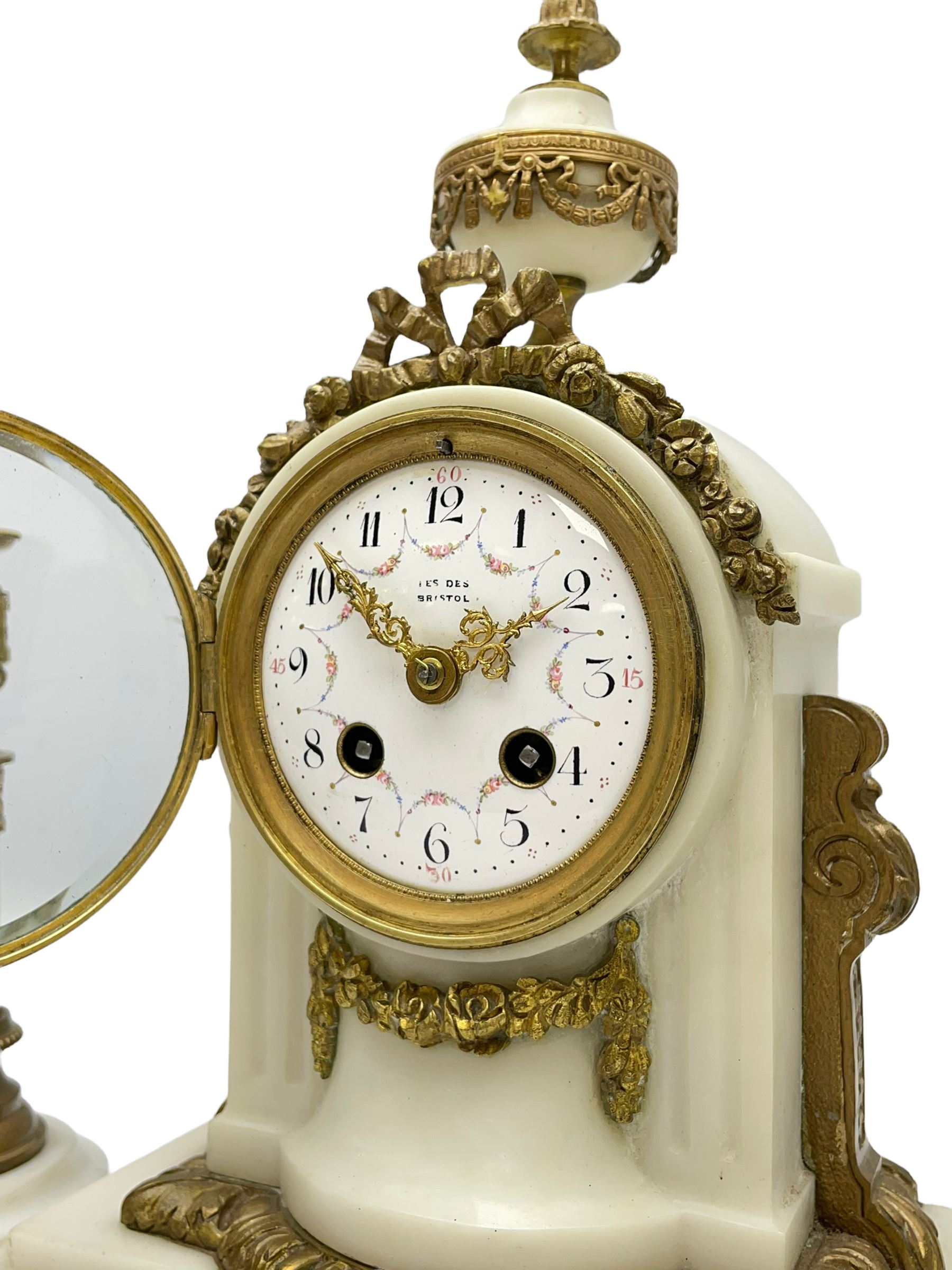 An early 20th century white marble mantle clock in a break front case with an arched top and finial, gilded scroll work to the sides and pierced foliate panels to the front, plinth raised on five bun feet, enamel dial with garland decoration, Arabic numerals, minute markers and Louis XV gilt hands, within a convex glazed bezel, eight-day Parisian striking movement striking the hours and half hours on a bell, with two conforming single light candlesticks. With Pendulum.

