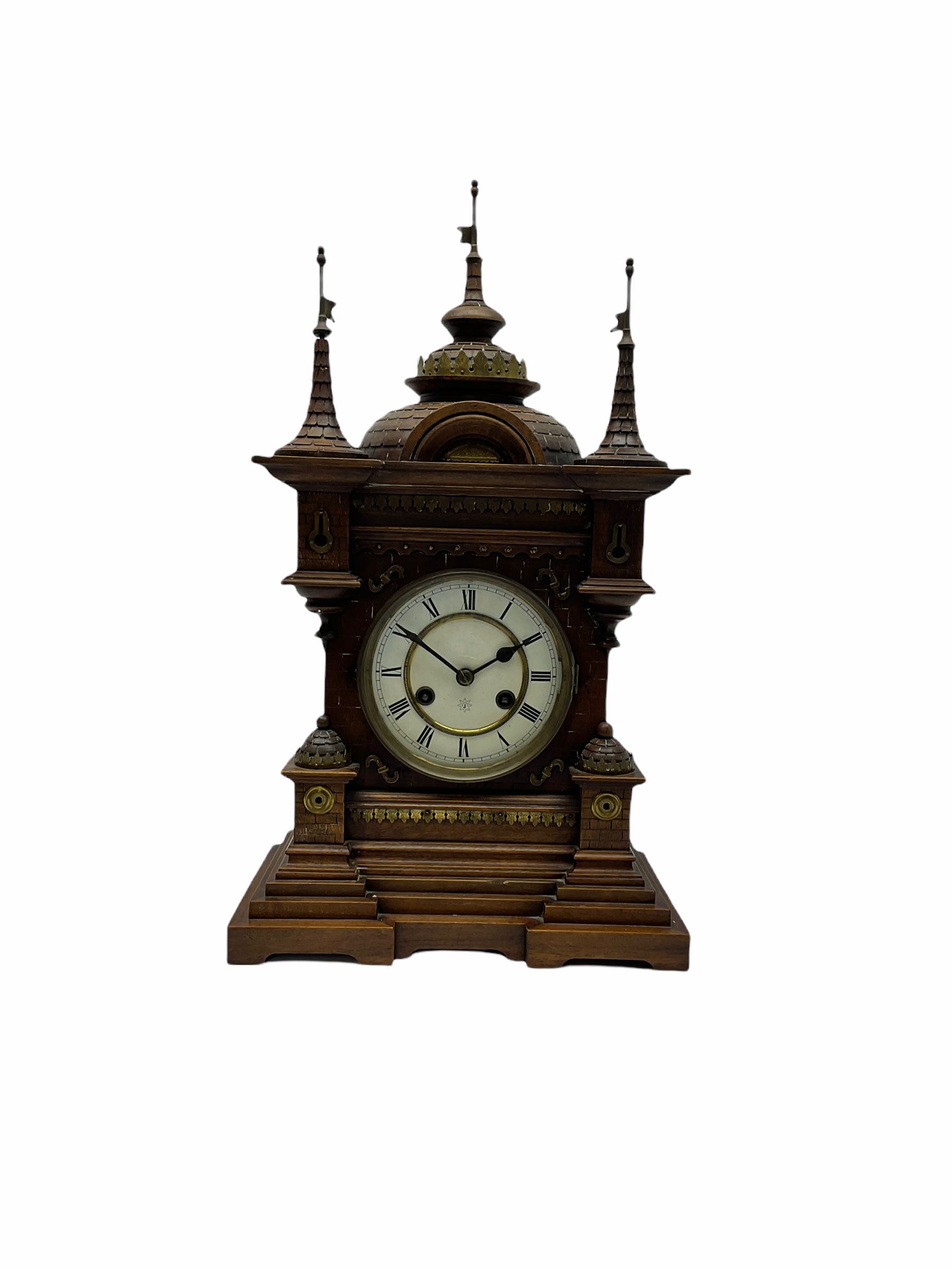 A late 19th century German Black Forrest mantle clock striking the hours and half hours on a coiled gong, two-piece white enamel dial inscribed with the six pointed Junghans logo, roman numerals and minute track, steel spade hands, spun brass bezel and glass, spring driven eight day movement with a recoil anchor escapement, carved oak case in fantasy form, multi-stepped plinth with incised brickwork effect to the sides and front of the case, with applied brass ornamentation, projecting turrets and gun ports, case surmounted with a pediment consisting of three distinctively carved oriental domes with roof tiles and guidon banner flags.
With pendulum.
