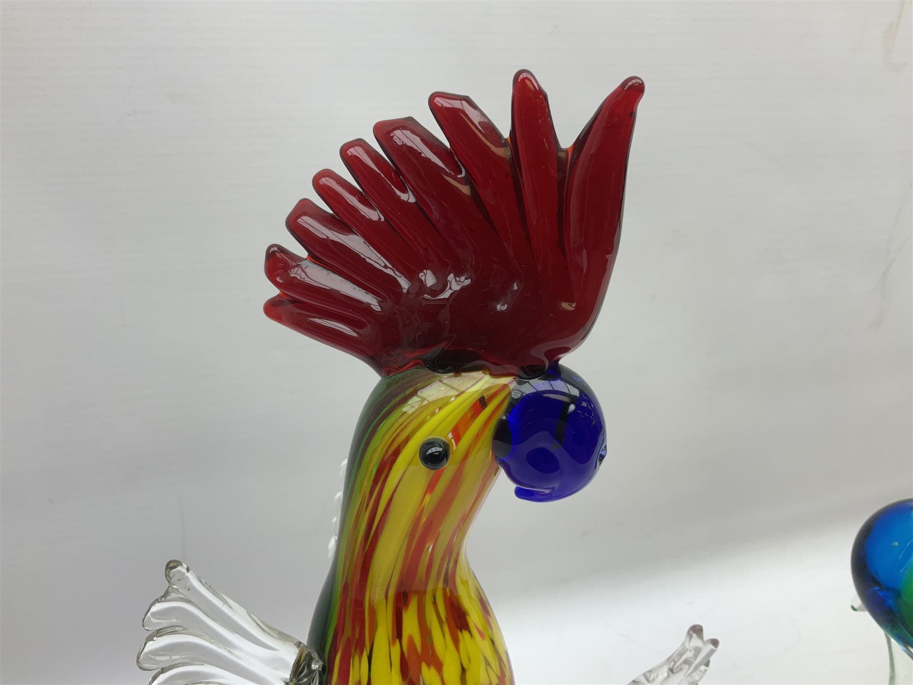 Three Murano style glass cockatoo parrots, the first with mottled yellow, red and orange body, clear outstretched wings, dark blue beak and red crest, raised upon clear stylised base, the second smaller parrot with merging blue and green body, and further with pink striped body, tallest H40cm