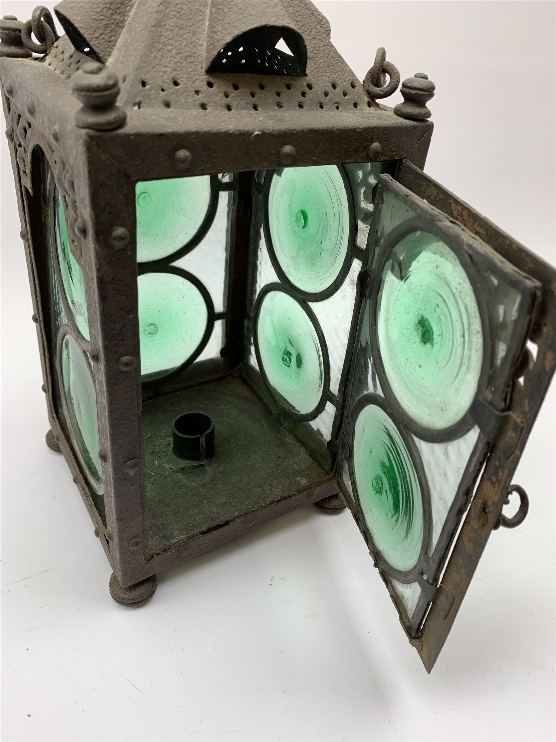 An Arts & Crafts wrought iron lantern, of square sided form with swing handle, and green and clear glass panels, raised upon four compressed bun feet, not including handle H30.5cm.