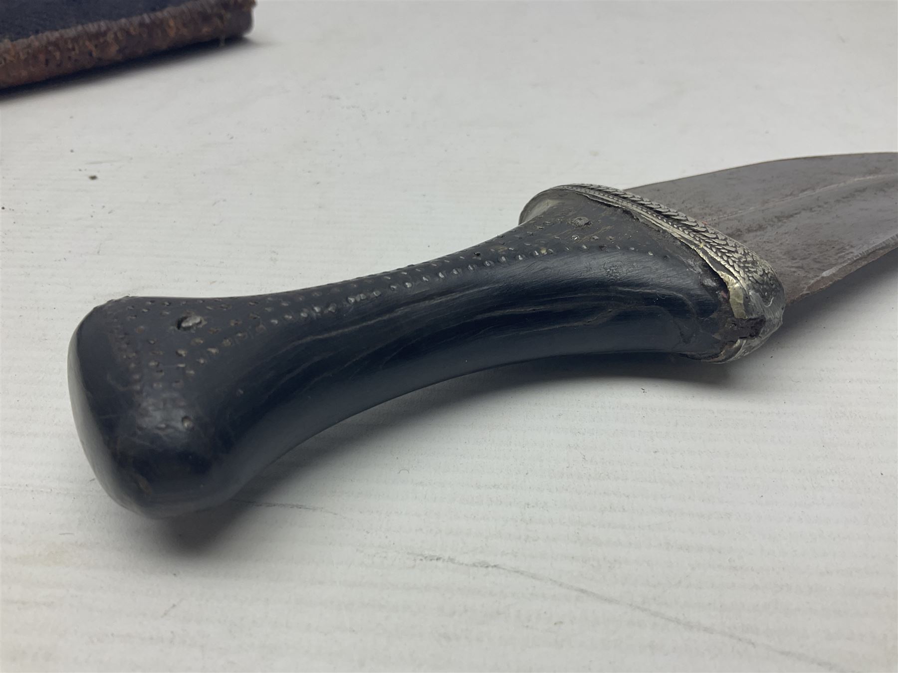 Eastern Jambiya, the 19cm double edge steel blade with raised medial ridge, waisted horn grip with pique work and white metal band; in stitched leather scabbard L33cm overall
