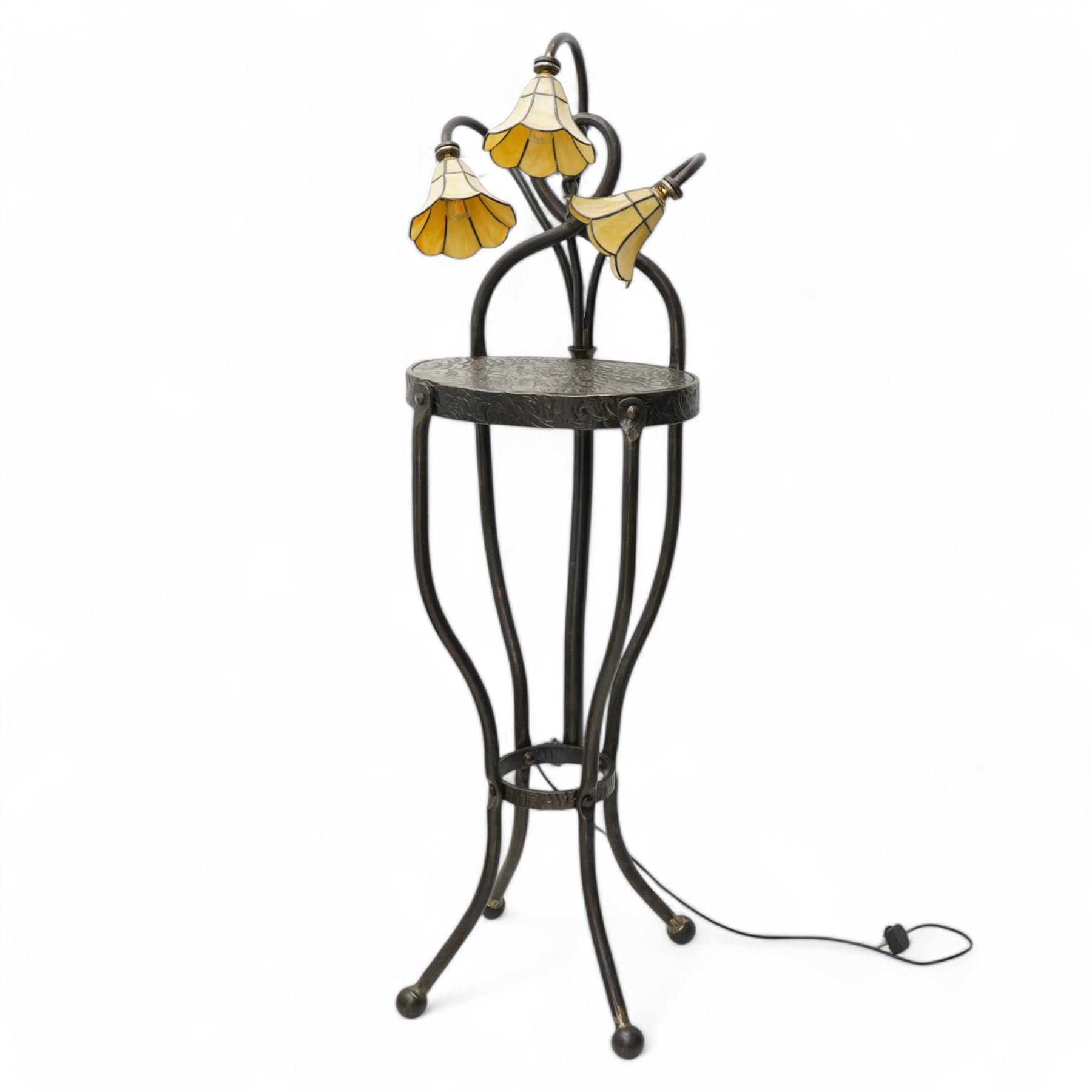 James Godbold - Art Nouveau blacksmith-made wrought iron floor standing lamp table, the round etched metal tabletop decorated with swirling foliage motifs, crowned by three Tiffany design amber glass shades mounted on curved iron stems, raised on four splayed supports united by circular stretcher and terminating in ball feet