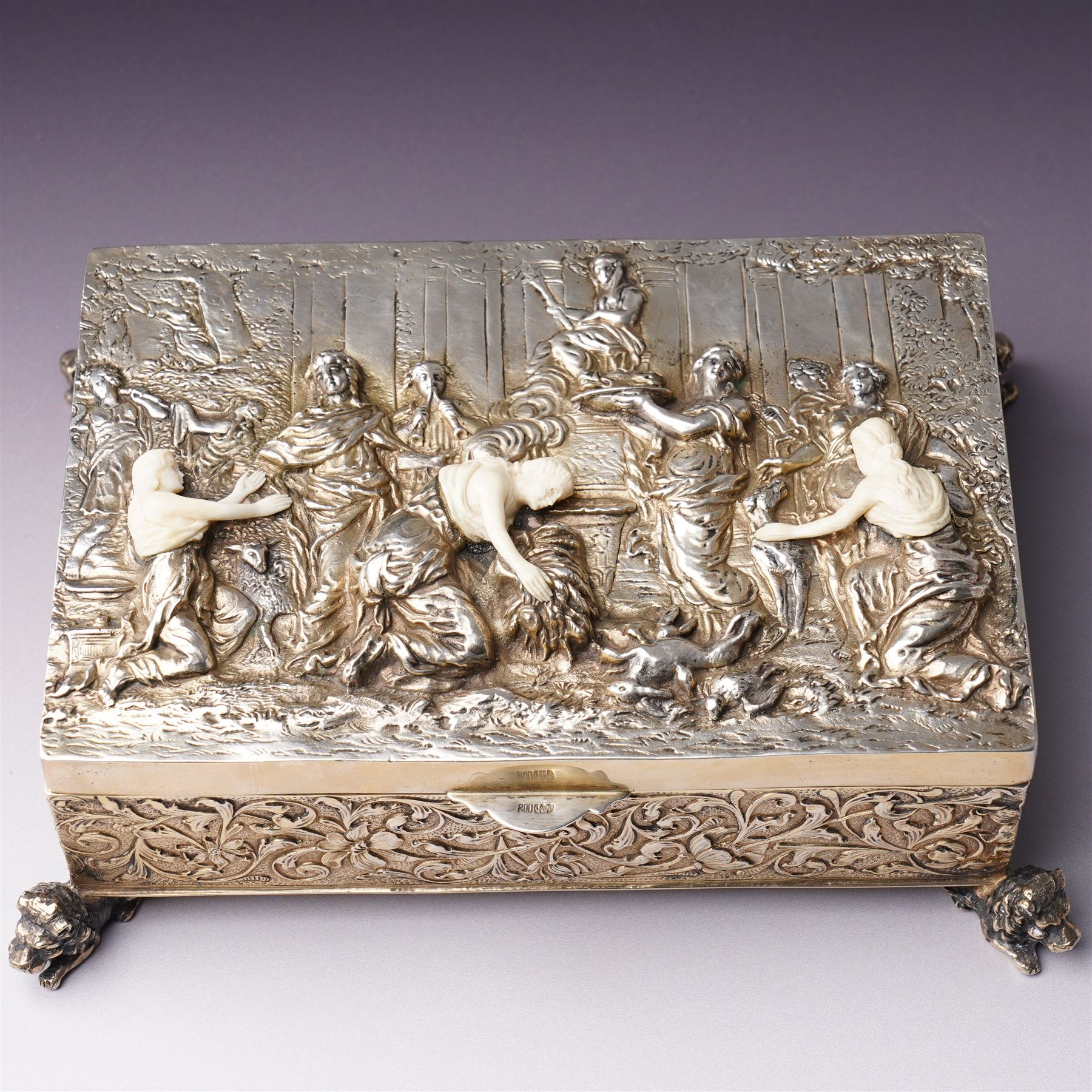 German 800 standard silver gilt and ivory mounted box, the hinged cover embossed and chased with an allegorical feasting scene, the sides with floral decoration, on four cast lion feet 21cm x 16cm x 8cm Maker Ludwig Neresheimer, Hanau circa 1900 This item has been registered for sale under Section 10 of the APHA Ivory Act