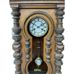 A late 19th century German wall clock in a mahogany case with a decorative carved pediment, full length shaped glazed door (glass missing) flanked by turned columns and pendant finials, with a two part enamel dial and steel gothic hands, eight-day striking movement striking the hours and half hours on a coiled gong, with a gridiron R/A pendulum. 

