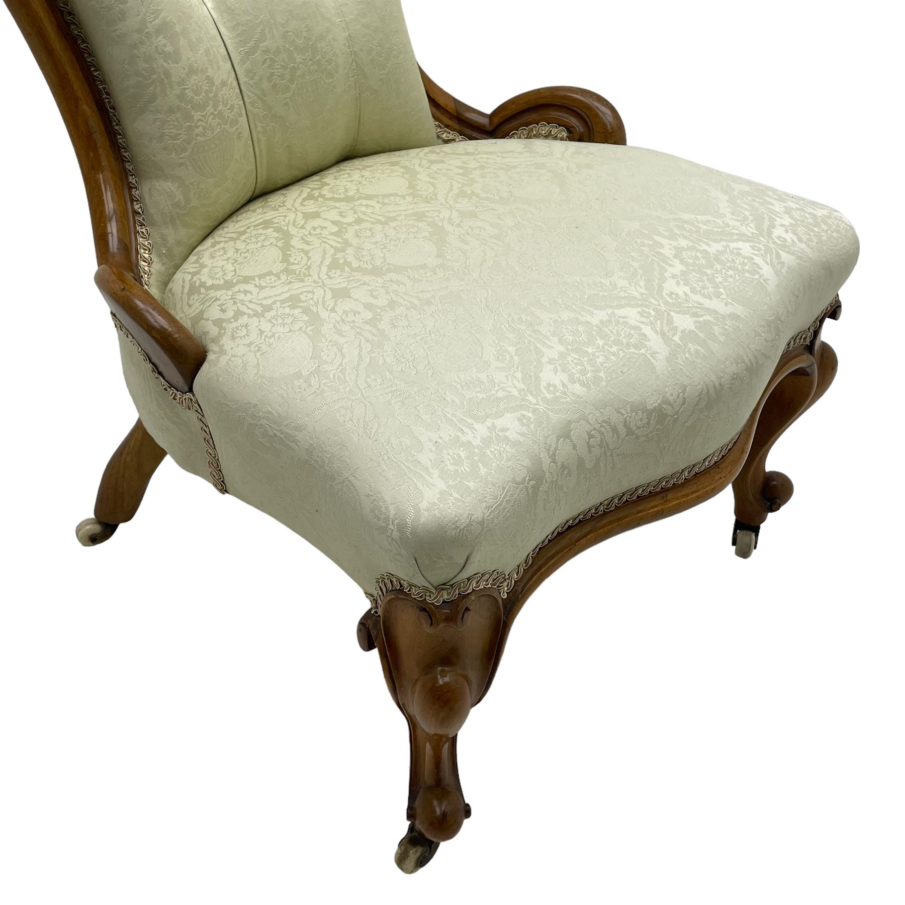 Victorian walnut framed nursing chair, the cresting rail decorated with carved applied flower heads and acanthus leaves, shaped back and sprung seat upholstered in buttoned ivory damask fabric, serpentine front over cartouche carved cabriole supports with ceramic castors (W57cm H92cm); with matched footstool, circular top over shaped moulded apron, on floral carved cabriole feet (W34cm) 