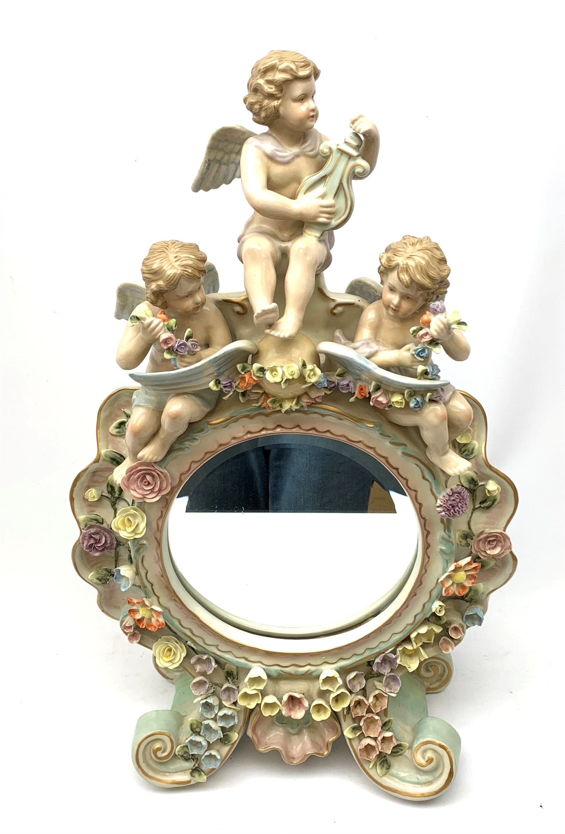  A late 19th century Sitzendorf porcelain mirror, the mirror plate of circular form set within a shaped surround encrusted with flowers and foliage, surmounted by three figures of cherubs and raised upon four scroll feet, with blue cross hatch mark beneath, H57cm.   