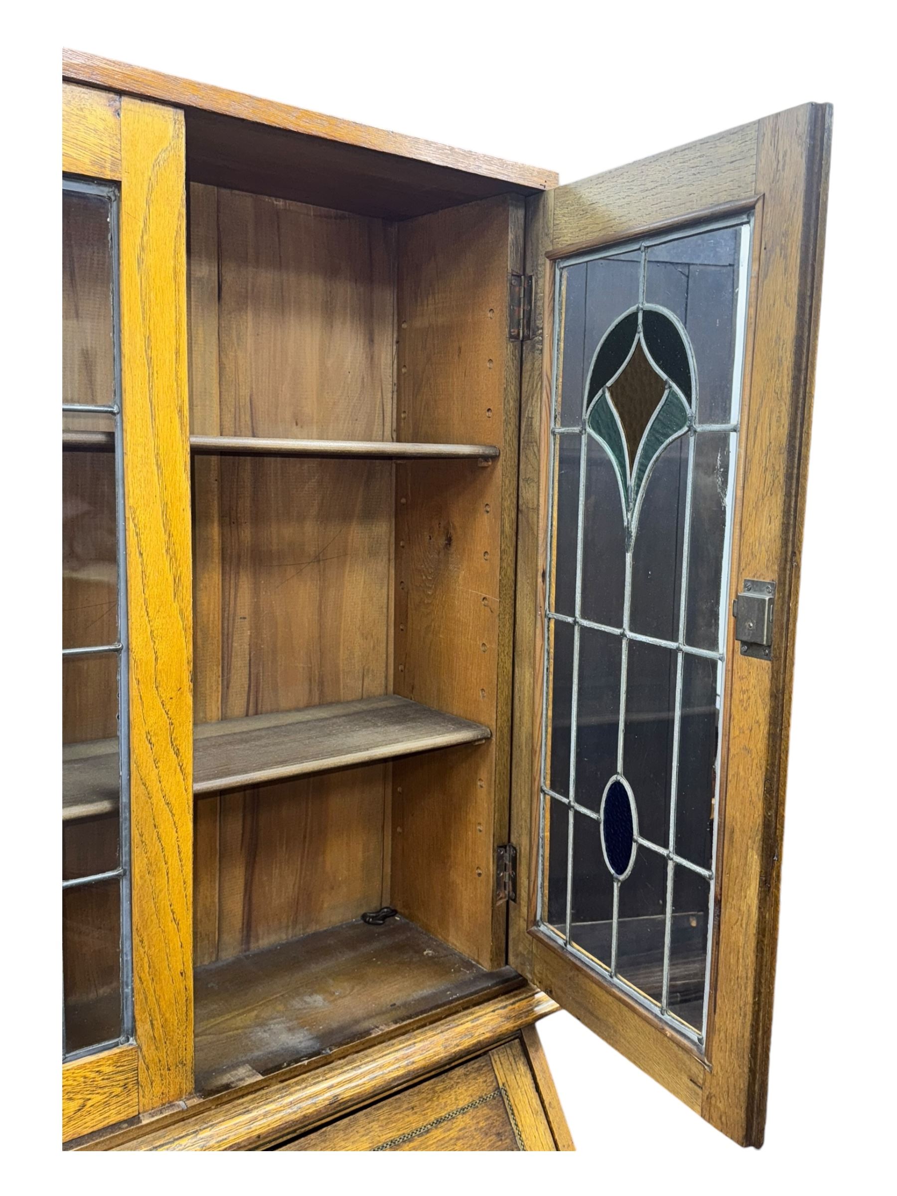 Early 20th century Arts and Crafts oak bureau bookcase, twin leaded and glazed stained glass doors enclosing two shelves, over bureau section with fall-front inlaid with sunburst motif, fitted interior of pigeonholes and shelves, on bracket feet