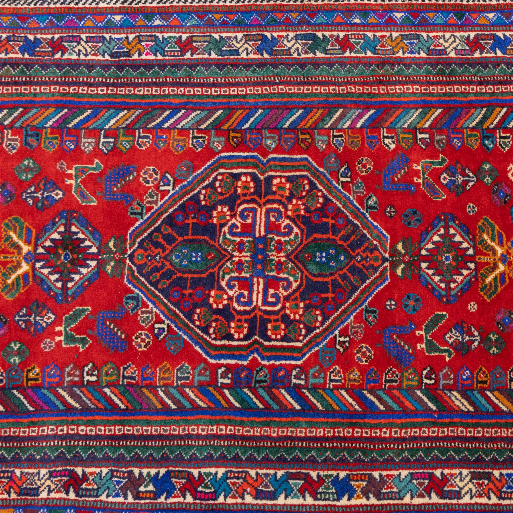 South West Persian Qashqai hand-knotted runner, madder red field  with central vertical succession of stepped lozenge medallions enclosing angular tribal devices and stylized floral motifs, principal medallion in deep indigo with ivory and sky blue highlights, field densely scattered with small geometric ornaments, hooked elements and zoomorphic forms, triangular indigo spandrels enclosed within multiple narrow guard stripes and principal border, with cotton fringe ends 