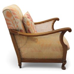 Pair of mid-20th century mahogany framed bergère armchairs, each with carved scroll arms and caned side panels, floral upholstered loose cushions, raised on cabriole front supports