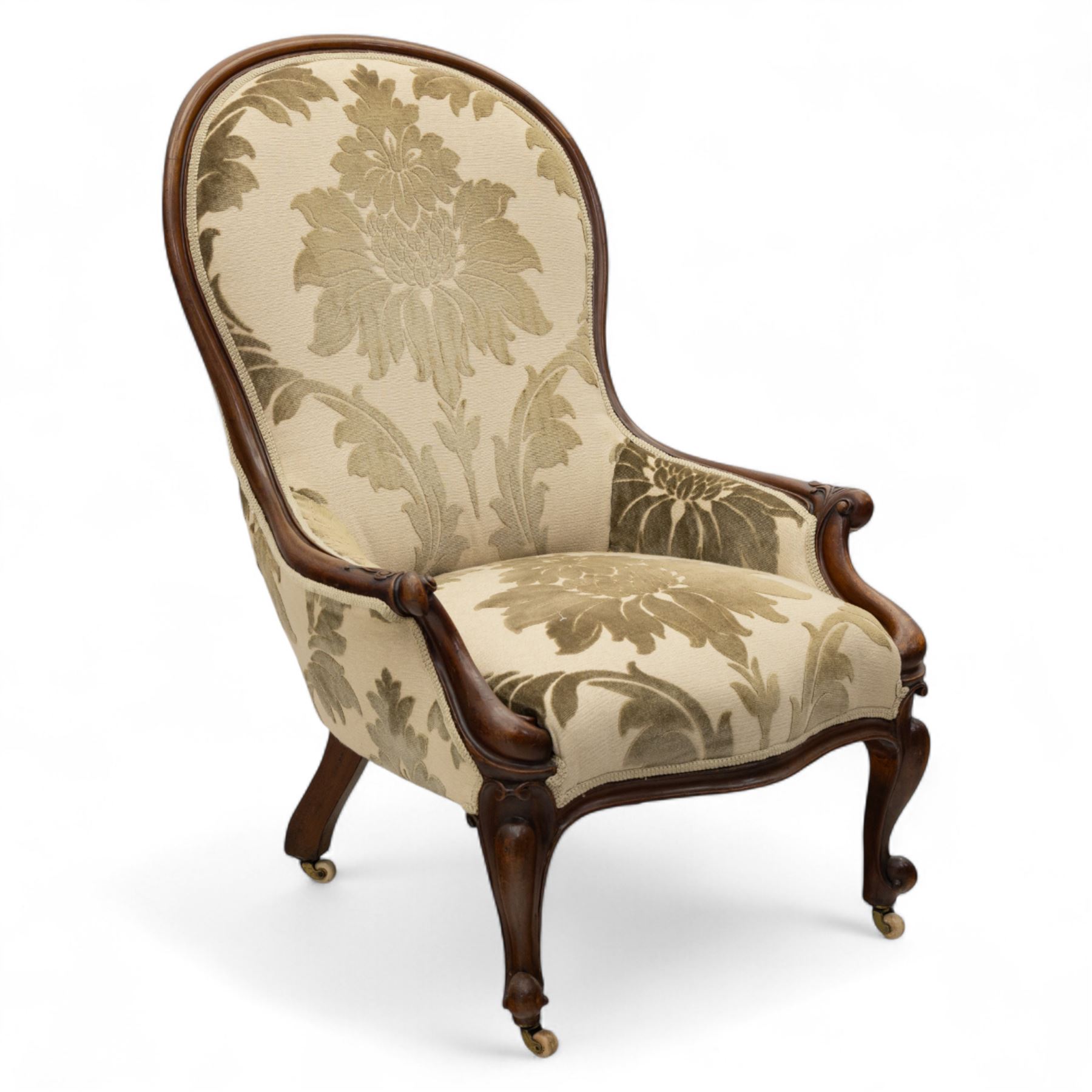 Victorian mahogany framed spoon back armchair, the shaped and padded back over serpentine seat upholstered in a floral damask pattern fabric within a moulded frame carved with scrolled arms, raised upon cabriole front legs with scroll feet on porcelain castors 