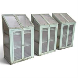 Three green-painted timber cold-frame greenhouses or growhouses, each with twin hinged sloping polycarbonate lids above a pair of panelled doors enclosing slatted shelves