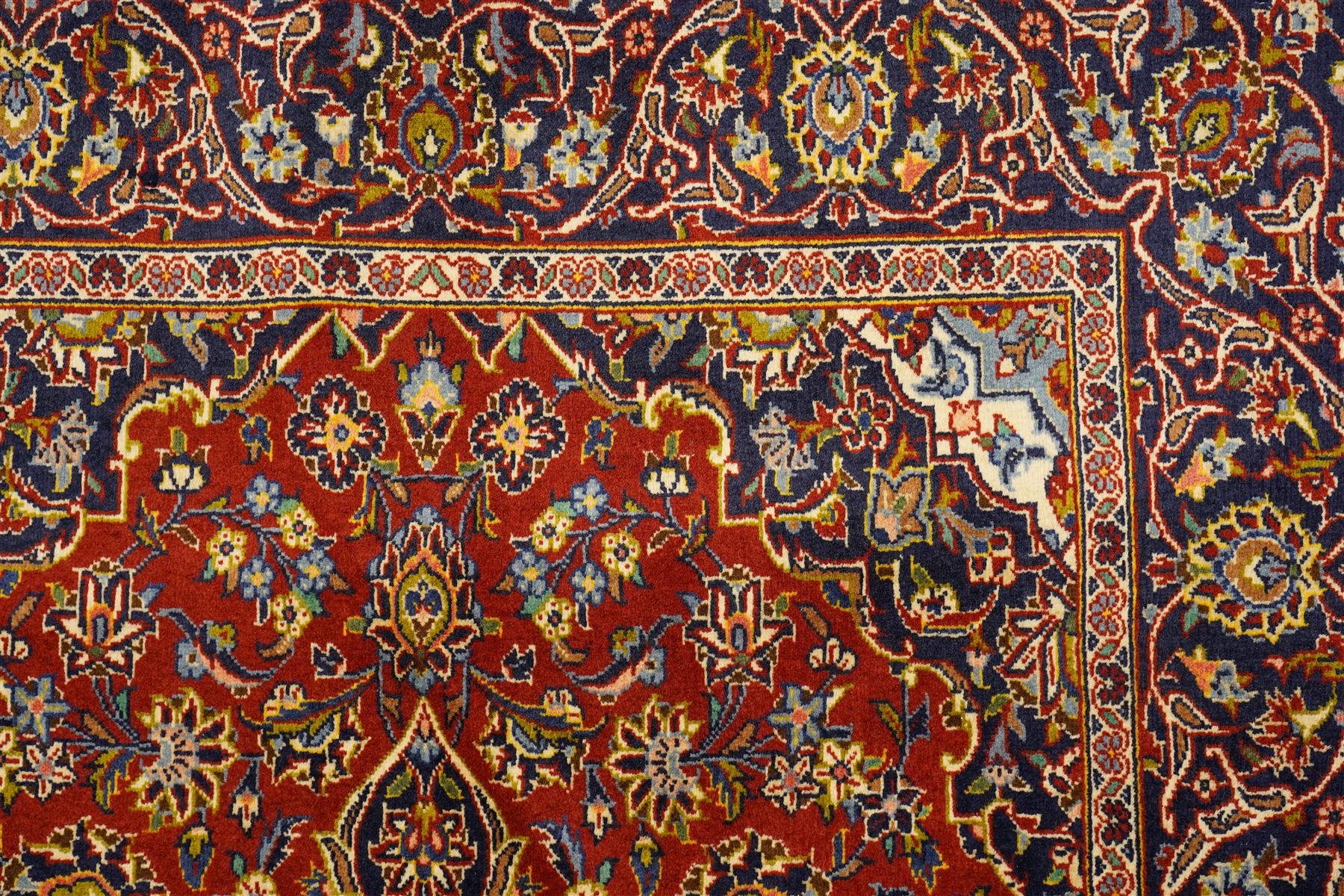 Central Persian Kashan hand-knotted rug, deep red field with central cusped ivory medallion with an all-over arrangement of scrolling floral vines, palmettes and flowering sprays within conforming spandrels, principal navy border with a continuous meandering vine of palmettes and stylized floral motifs, enclosed by multiple narrow guard borders, with cotton fringe ends 