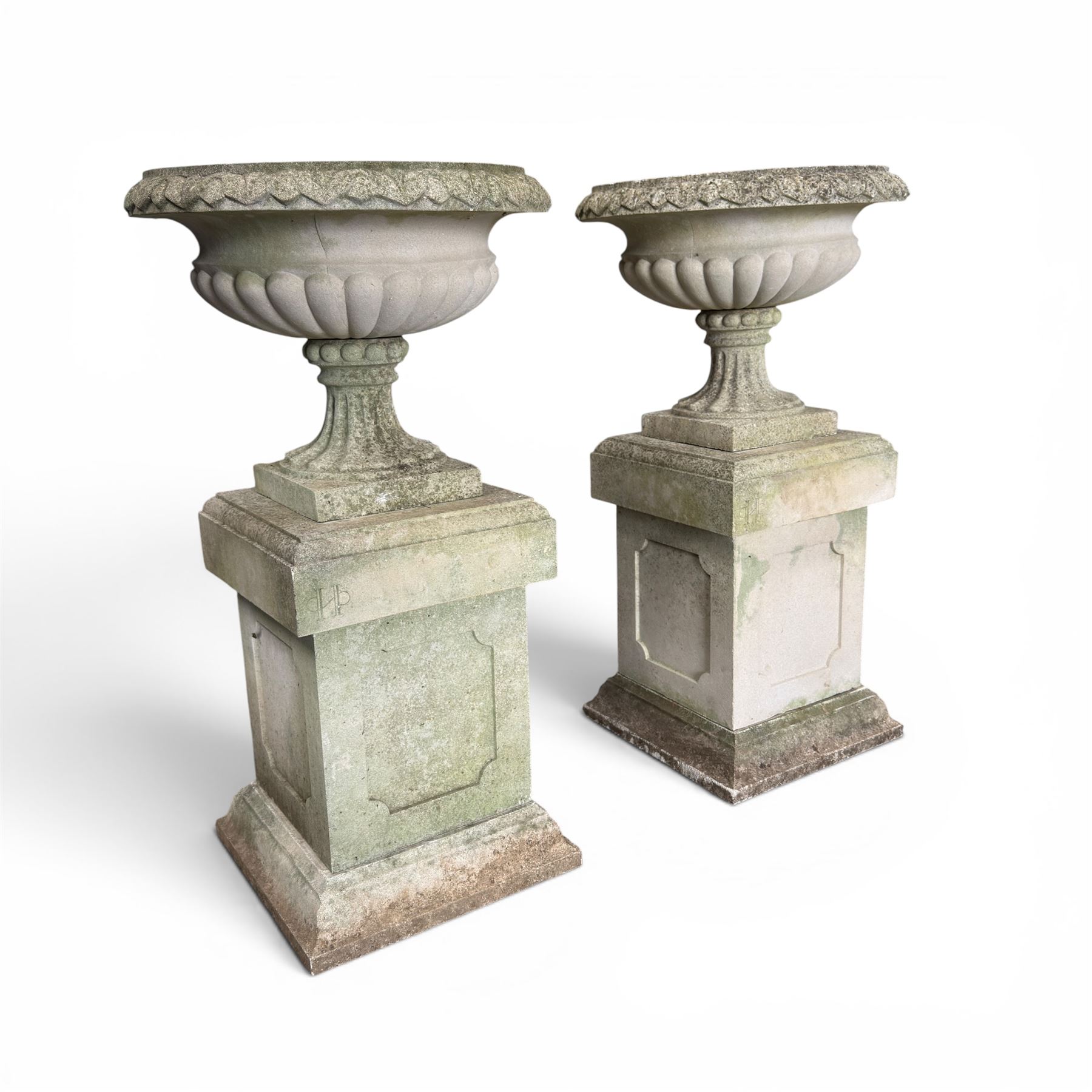 Pair of weathered cast stone Neoclassical design garden Kylix shape urns, foliate moulded rim over a gadrooned underbelly, the scalloped socle over a rectangular pedestal with faux-panelled sides, stamped with stylised ‘H’ monogram