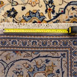 Central Persian Kashan hand-knotted carpet, ivory ground with central stepped floral medallion surrounded by scrolling vine work, palmettes, and stylized blossoms, field enclosed by a multi-border arrangement, wide main border decorated with repeating floral cartouches and foliate motifs, flanked by narrow guard stripes, with cotton fringe ends