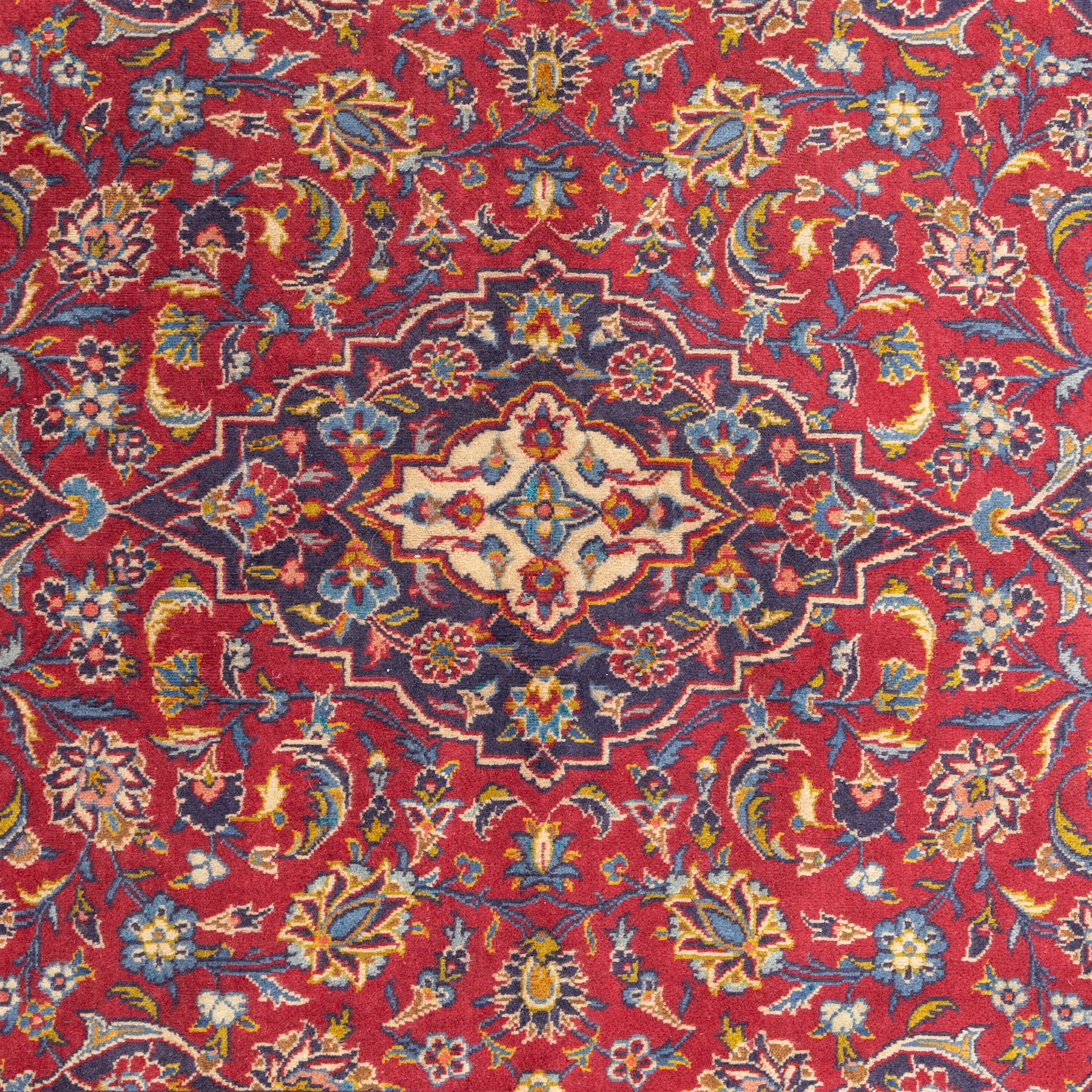 Central Persian Kashan hand-knotted rug, crimson field, profusely decorated with scrolling vines and palmettes, floral sprays around an ivory and indigo lobbed medallion enclosed by spandrels within an indigo border of arabesques between multiple guard stripes  