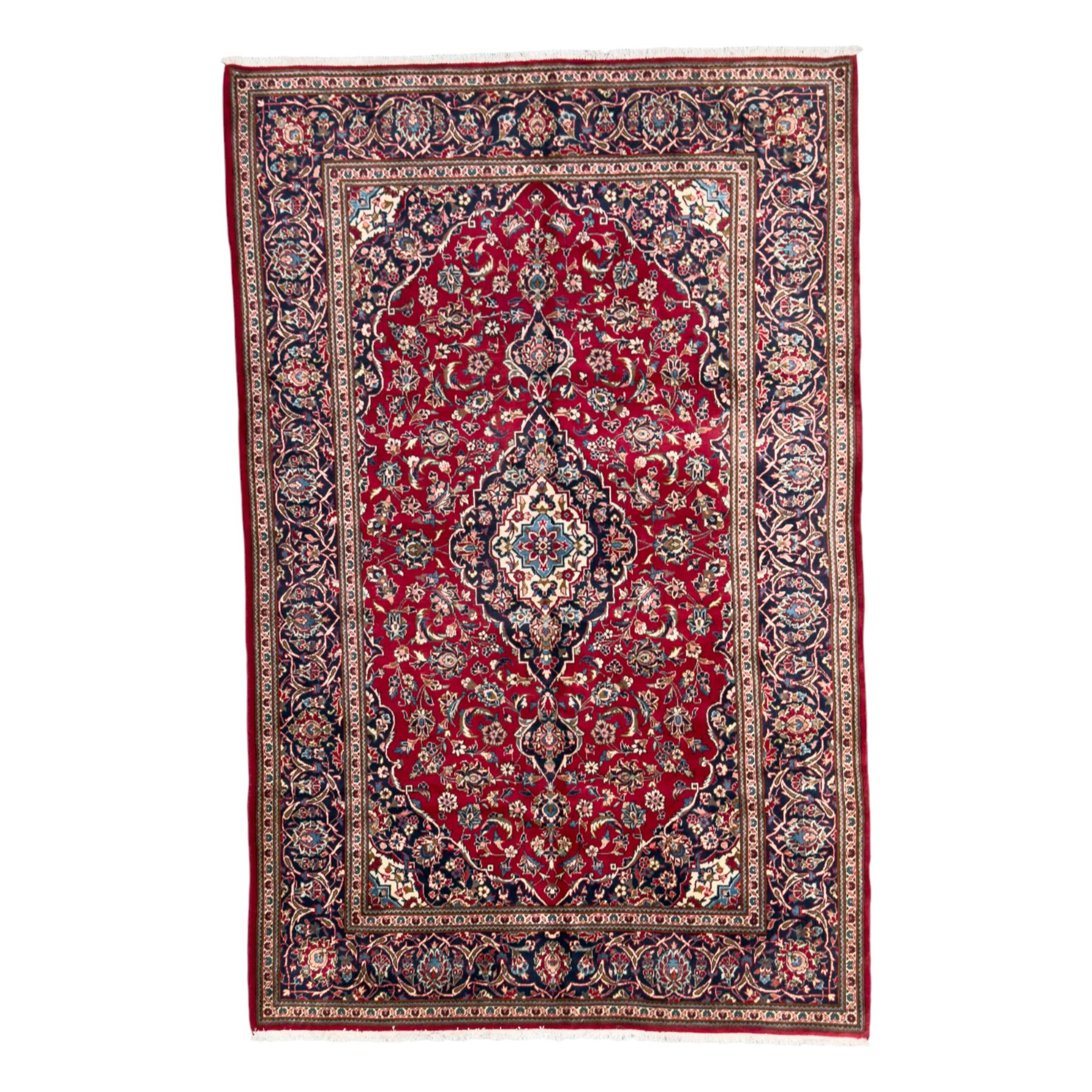 Central Persian Kashan hand-knotted carpet, the deep red ground with a central lozenge medallion in ivory and navy, surrounded by dense scrolling floral and vine motifs, navy spandrels and a wide border with stylised palmettes and flowerheads enclosed by multiple guard stripes