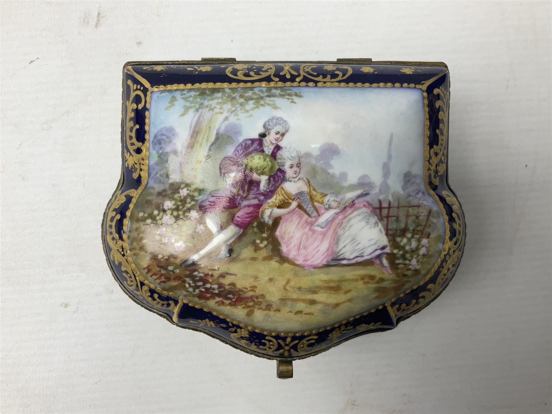 Sevres style late 19th/early 20th century trinket box decorated in the rococo style, the shaped cover painted with an 18th century courting couple in a garden scene opening to reveal the interior painted with floral sprays, the conforming body with a painted panel of floral decoration, all reserved upon cobalt blue ground with foliate gilt borders, with interlaced L's mark beneath, L12cm H6cm