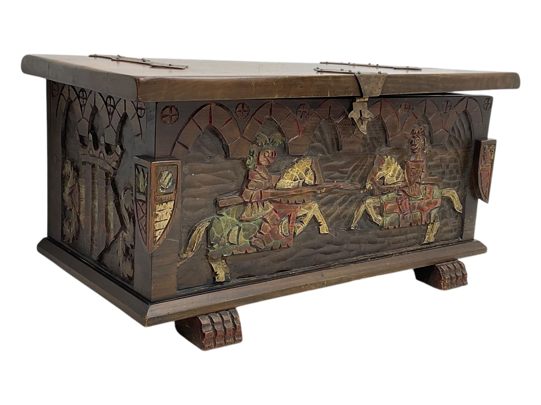 Mid-20th century carved hardwood chest, the rectangular hinged top with decorative strap hinges, the front and sides deeply carved with medieval-style jousting knights, heraldic shields, and architectural elements, raised on shaped block feet