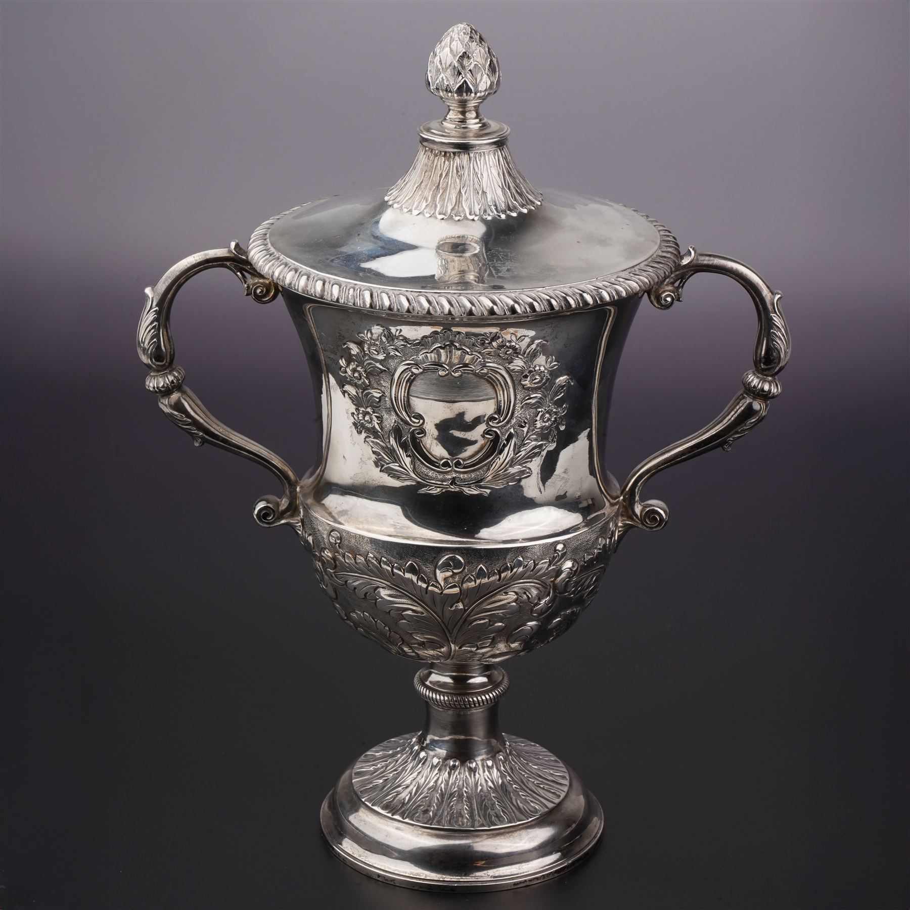 George III silver two handled campana shape cup and cover with pineapple lift, the body with cartouche surrounded by flowers above leaves and scrolls on a pedestal foot with acanthus leaf decoration H29cm London 1764, makers marks rubbed