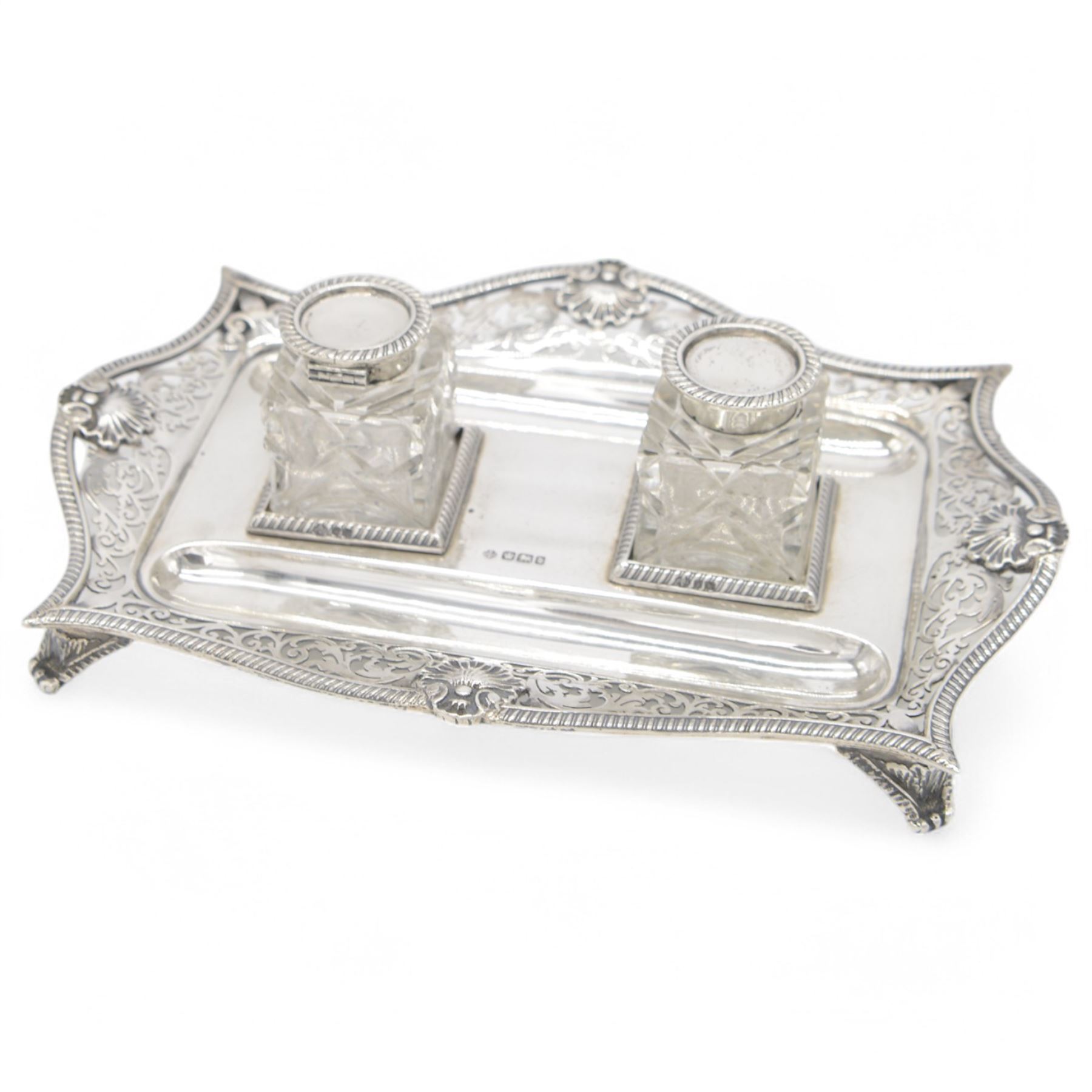 Late Victorian silver desk stand, of rectangular form, with shaped and pierced, oblique gadrooned rim, upon four paw feet, the central tray with two pen holders and two reserves holding cut glass silver topped inkwells, hallmarked William Hutton & Sons Ltd, Sheffield 1900, tray W23cm