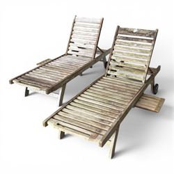 Pair of teak garden sun loungers, with slatted adjustable backrests, integrated wheels and sliding pull-out side trays, weathered natural finish.