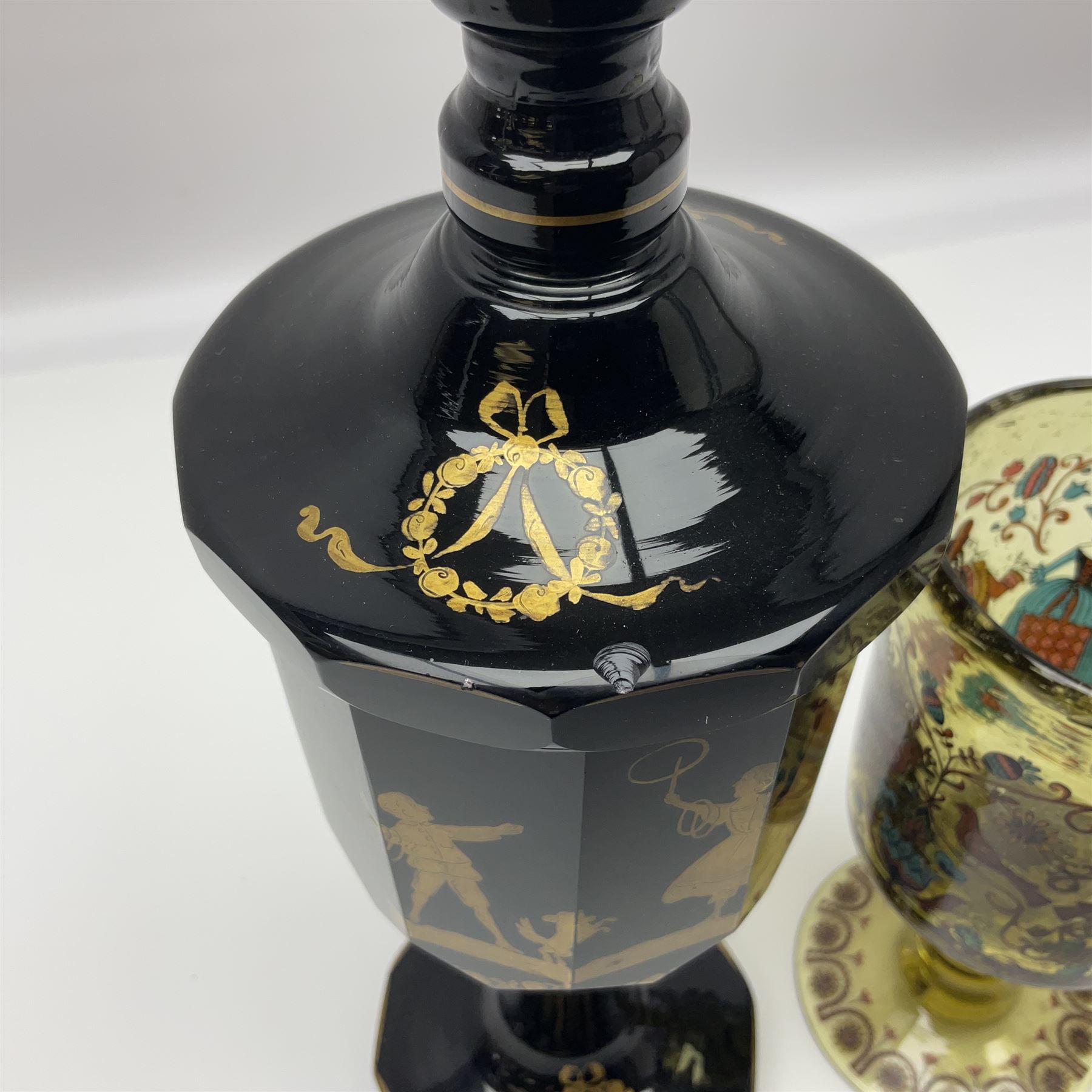 19th Century Bohemian amethyst glass lidded goblet, the bowl of faceted form, gilded with three children in period dress playing games, upon a knopped stem and conforming faceted foot, together with an art nouveau green glass goblet, with gilded rim, upon clear trumpet stem, and a hand blown wine glass, transfer printed with birds and three figures in period dress, tallest H29cm