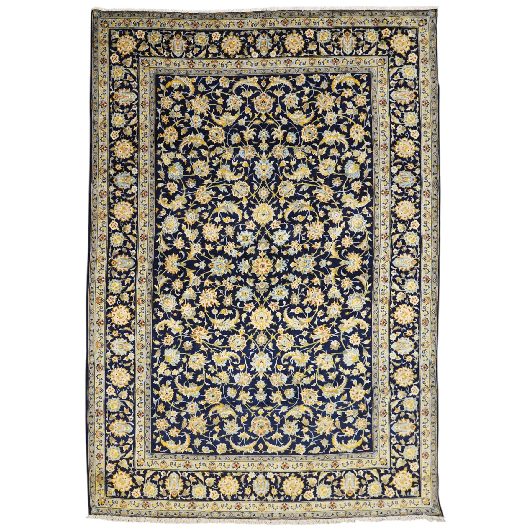 Central Persian Kashan hand-knotted carpet, deep indigo field with a densely all-over arrangement of scrolling floral vines, palmettes, and flowering sprays, in soft ivory, pale blue, and warm gold tones, principal ivory border with a continuous meandering vine of stylized palmettes, and floral motifs, flanked by multiple narrow guard borders, with cotton fringe ends 