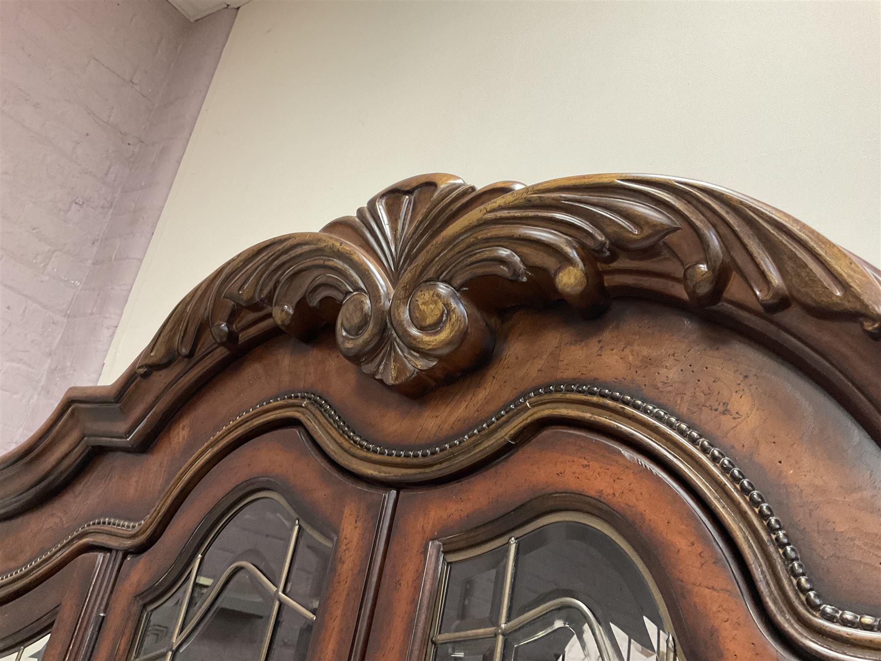 Kevin Charles American walnut display cabinet, illuminated interior, shell carved pediment above four bevel edge doors enclosing six glazed shelves above one central long drawer flanked by two short drawers, above three cupboards, carved bracket supports 