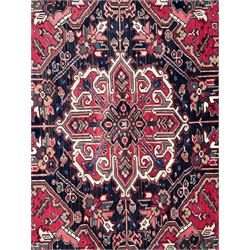 Persian Heriz red ground rug, central scrolled medallion on dark ground decorated with floral motifs, the surrounding field decorated with stylised plant motifs and smaller geometric symbols, guarded border with repeating pattern punctuated with rosettes and flower heads 