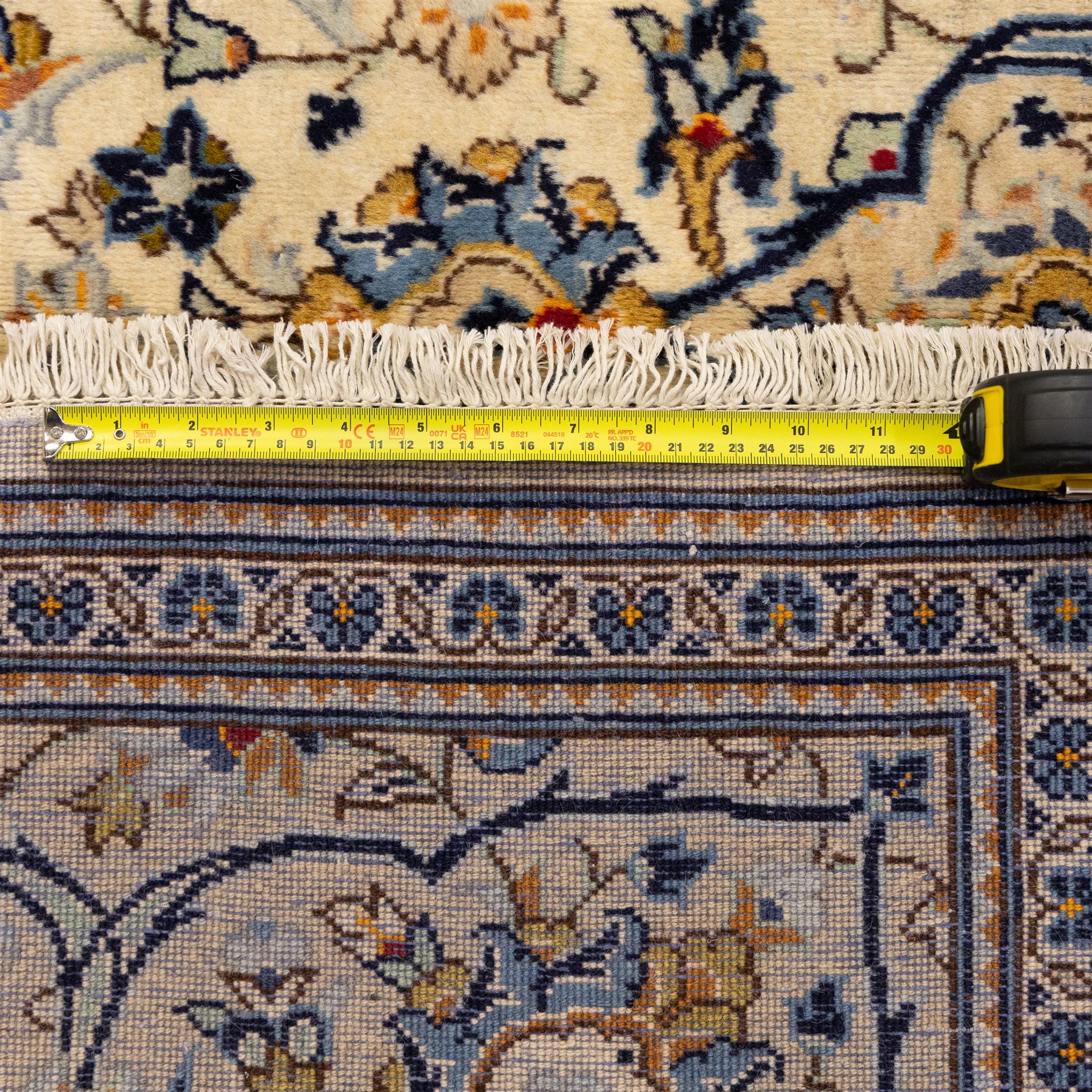 Central Persian Kashan hand-knotted carpet, ivory ground with central stepped floral medallion surrounded by scrolling vine work, palmettes, and stylized blossoms, field enclosed by a multi-border arrangement, wide main border decorated with repeating floral cartouches and foliate motifs, flanked by narrow guard stripes, with cotton fringe ends