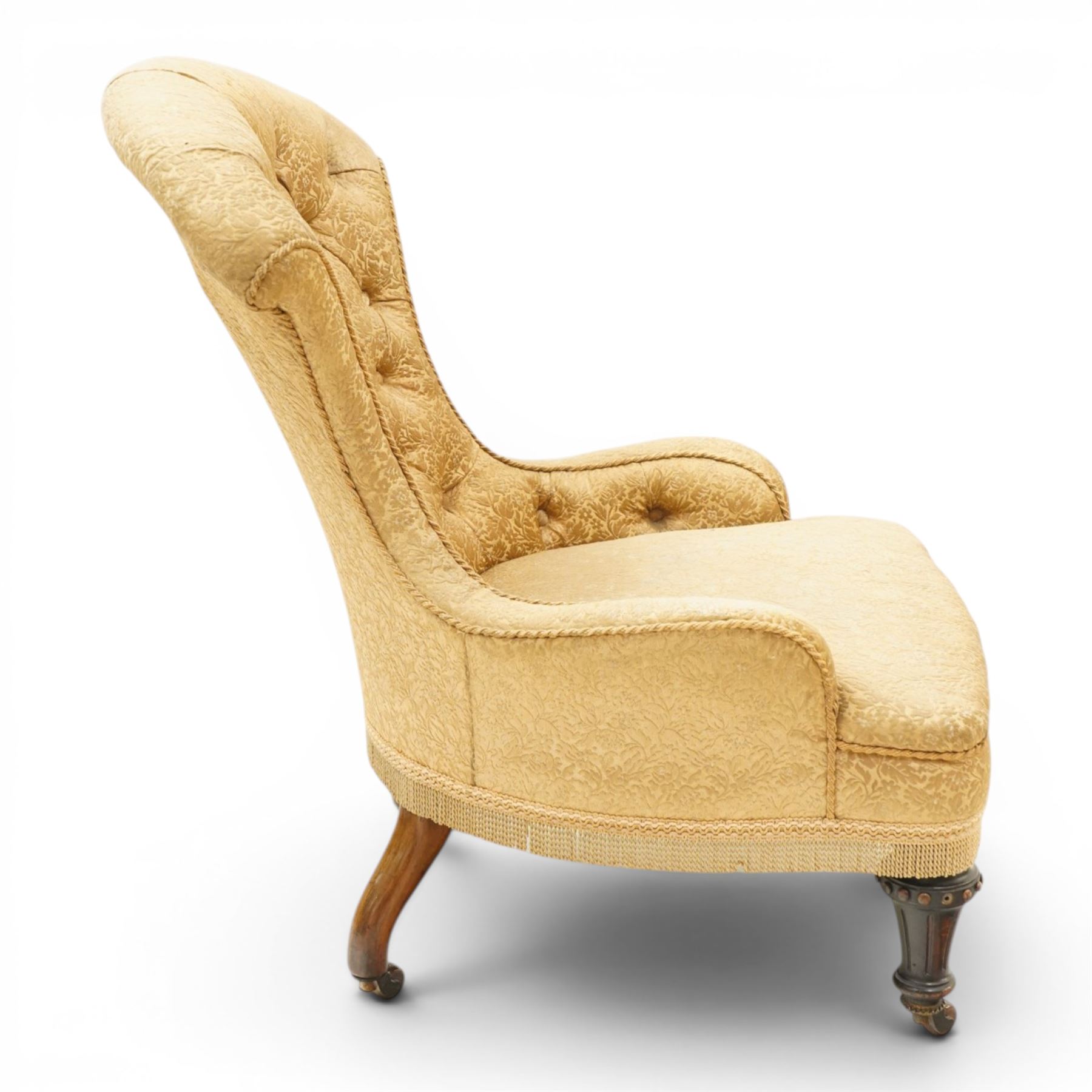 Victorian Aesthetic Movement walnut framed nursing chair, the fan back and sprung seat upholstered in buttoned foliate patterned gold damask fabric with fringing, raised on beaded tapering fluted supports with ceramic castors