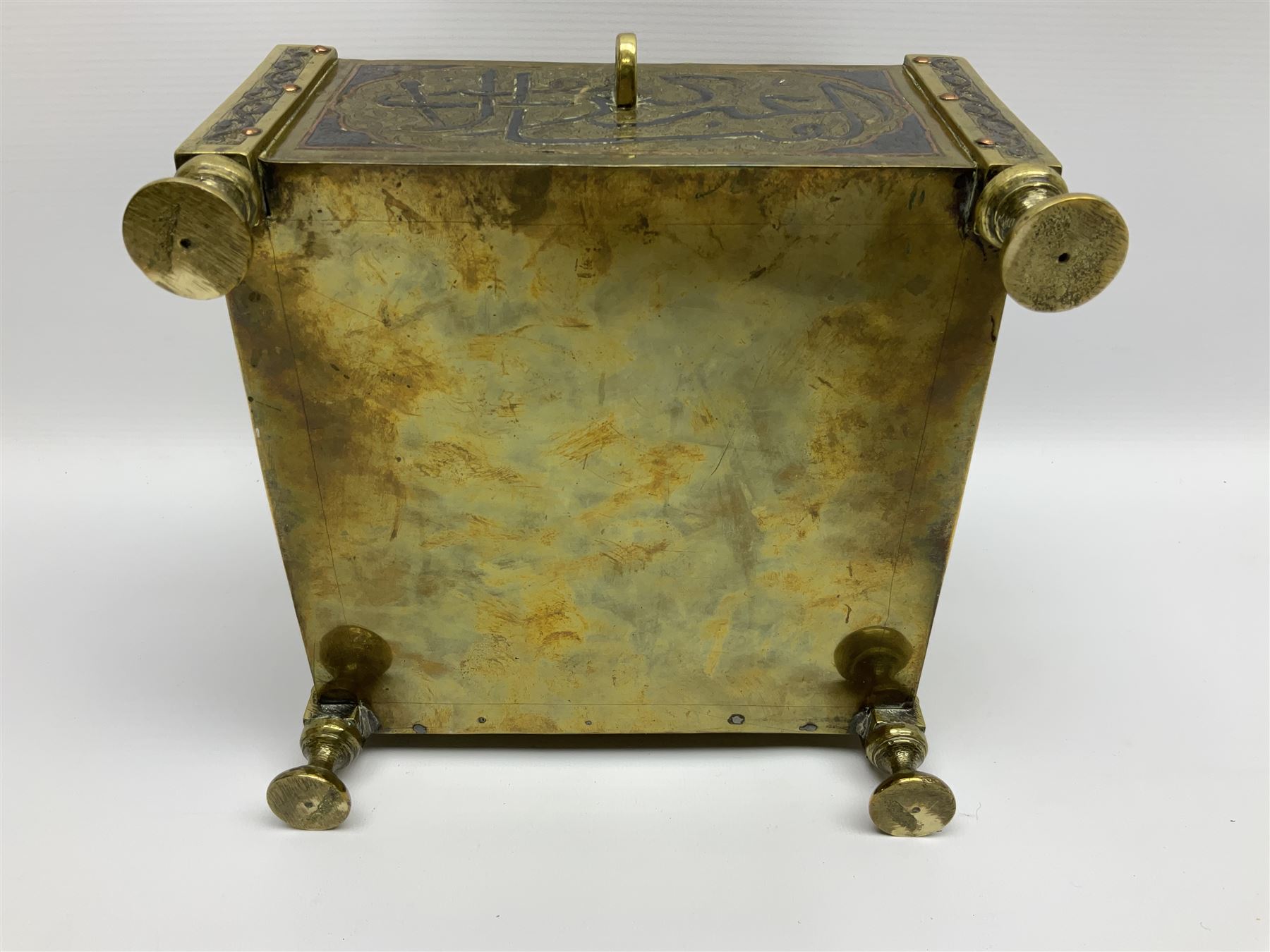Late 19th century Damascus Cairoware Quran box, of square form with canted hinged cover, upon four circular pad feet, the brass exterior inlaid with silver and copper script and decorative detail, opening to reveal a marquetry lined interior, H15.5cm W19cm D19cm