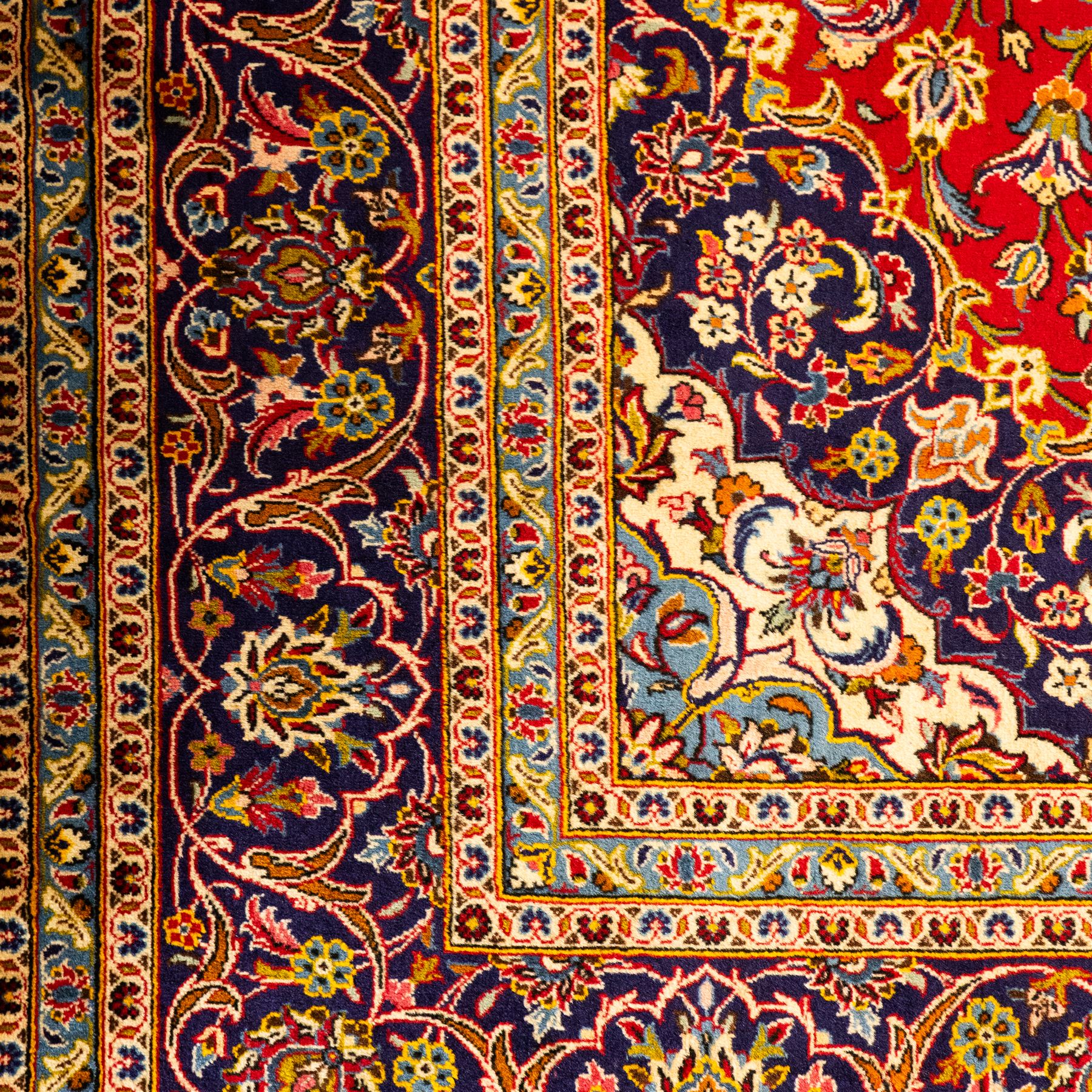Central Persian Kashan hand-knotted carpet, central field in a ruby red ground with an indigo medallion with pendant motifs, enclosing scrolling arabesques, palmettes, and floral sprays in shades of indigo, turquoise, ivory, and gold, framed by indigo spandrels and a broad floral border between multiple guard stripes 