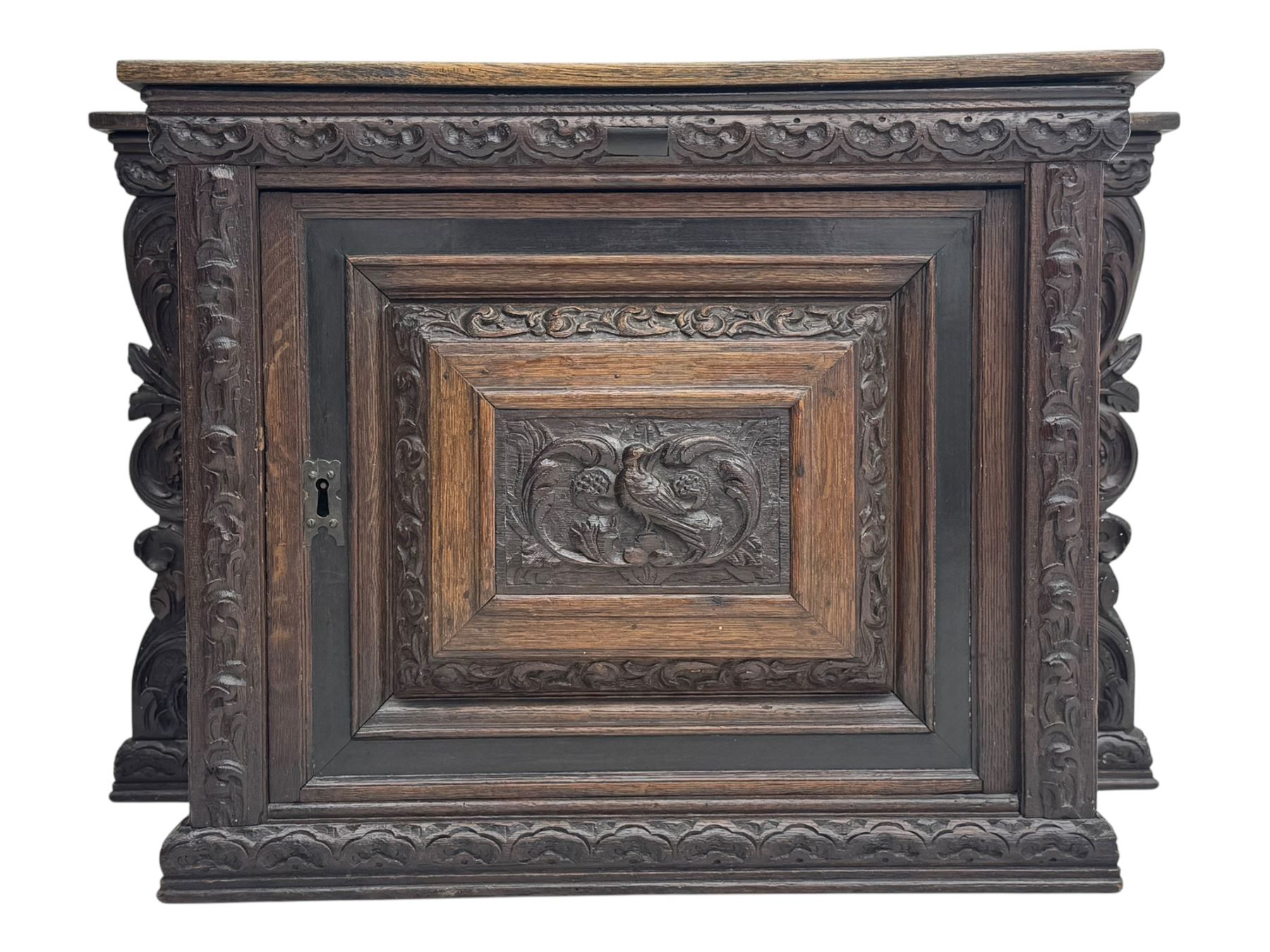 18th century and later carved oak cupboard, enclosed by panelled door carved with central bird motif and scrolled leafage with an outer ebony band, the frieze and uprights carved with repeating lunette and floral designs, flanked by scrolled foliate carved upright brackets