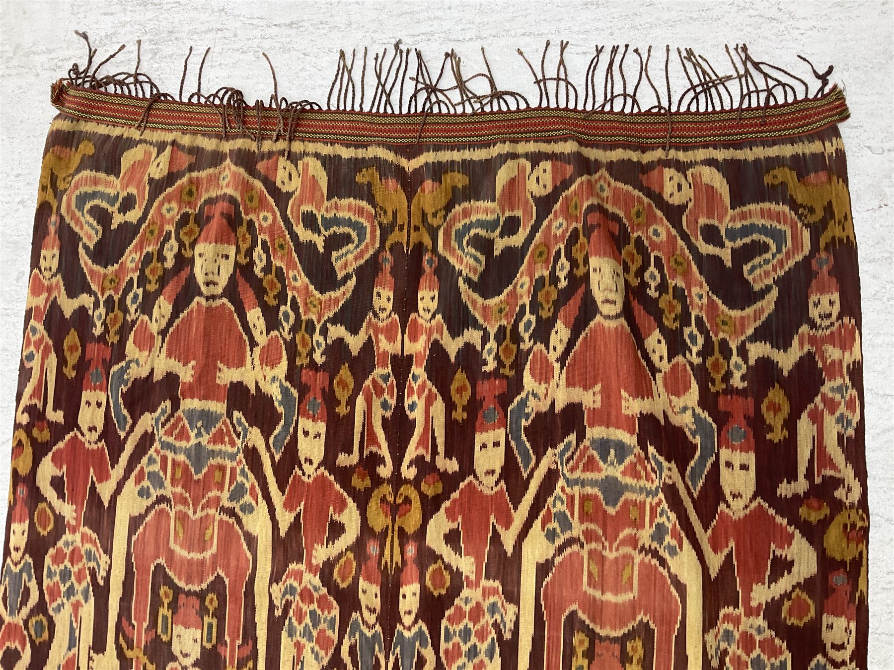 Ikat Man’s Cloth (Hinggi Kombu), East Sumba, Indonesia, woven in red, blue and yellow-brown warp ikat, constructed of two mirroring long panels, the motifs arranged in four bands depicting warrior figures astride rearing horses, high-ranking figures and guardians within geometric borders, on carved wooden wall hanger, L295cm
