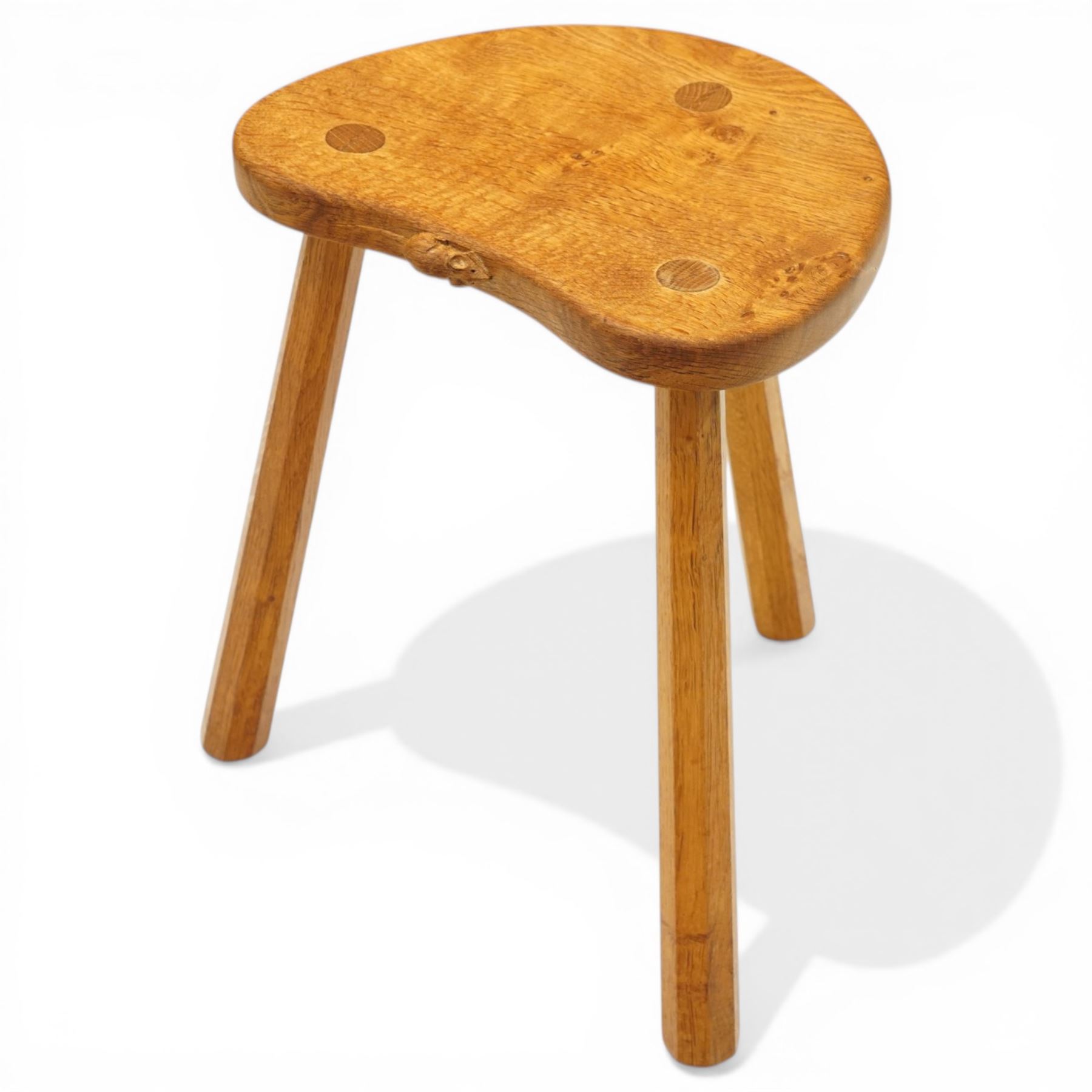 Mouseman - oak three-legged stool, dished kidney-shaped seat with burr inclusions, raised on three octagonal splayed supports, with carved mouse signature, by the workshop of Robert Thompson of Kilburn
