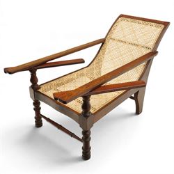 20th century teak framed plantation chair, plain cresting rail over cane back and seat, fitted with hinged extending leg rests, raised on turned front-supports united by ring-turned stretcher