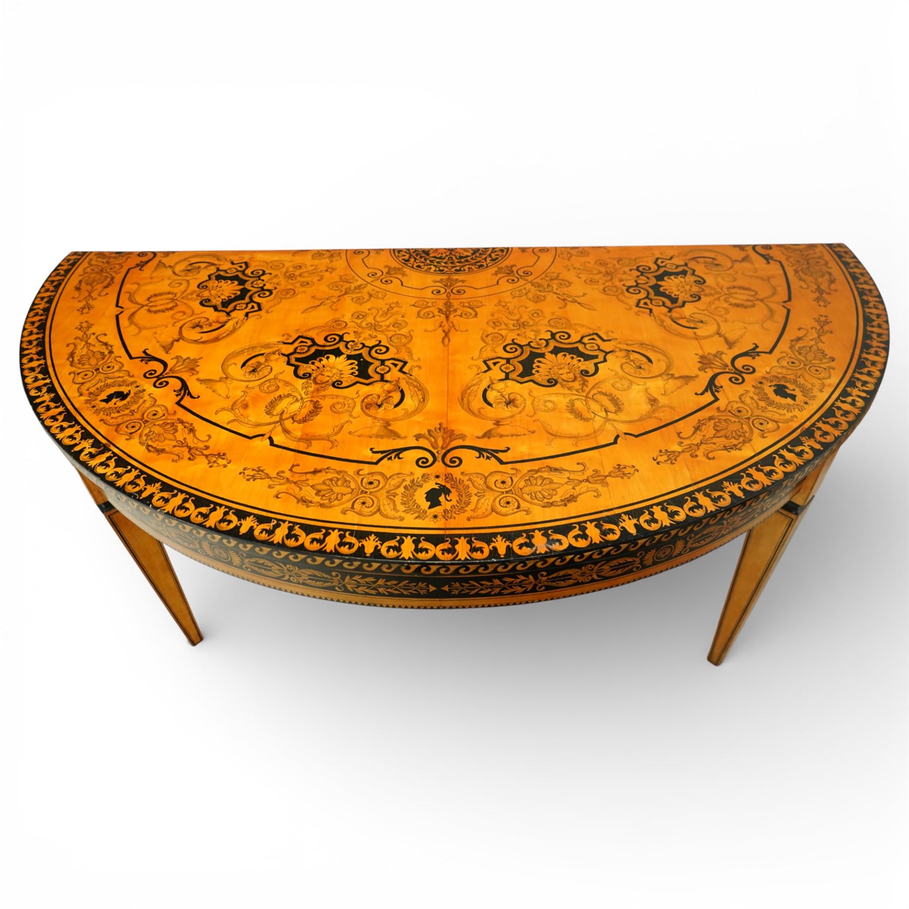 20th-Century Louis XVI-Style satinwood and penwork circular table or pair of console tables, the circular quarter-veneered top decorated in penwork with a central classical mask head motif within scrolling foliate and anthemion ornament on a contrasting black ground, above a conforming frieze decorated with Greek key-type scrolls. Raised on square tapering legs, each with an upper block panel decorated with compass-star motifs.