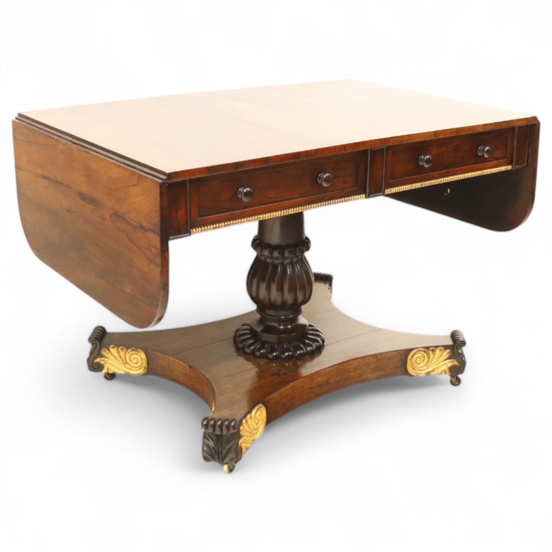 Regency rosewood sofa table, rectangular top with variegated rosewood veneers, hinged D-shaped leaves, over a set of dummy drawers to the back and full-size drawers to the front, with turned knob handles and gilt beaded moulding on frieze above elegant turned, tapering, and fluted, pedestal with a well-carved collar, supported on a large, tripartite concave platform base terminating in richly carved lion paw feet with gilded acanthus leaf carving, on concealed brass castors, circa 1815, length extended 161cm 
