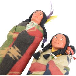 Two Skookum Native American Indian dolls with foot label, with painted composition heads, side glancing eyes, black hair wig, stuffed rigid body, original clothes, the male with feather headdress, the female with baby on back, H32cm