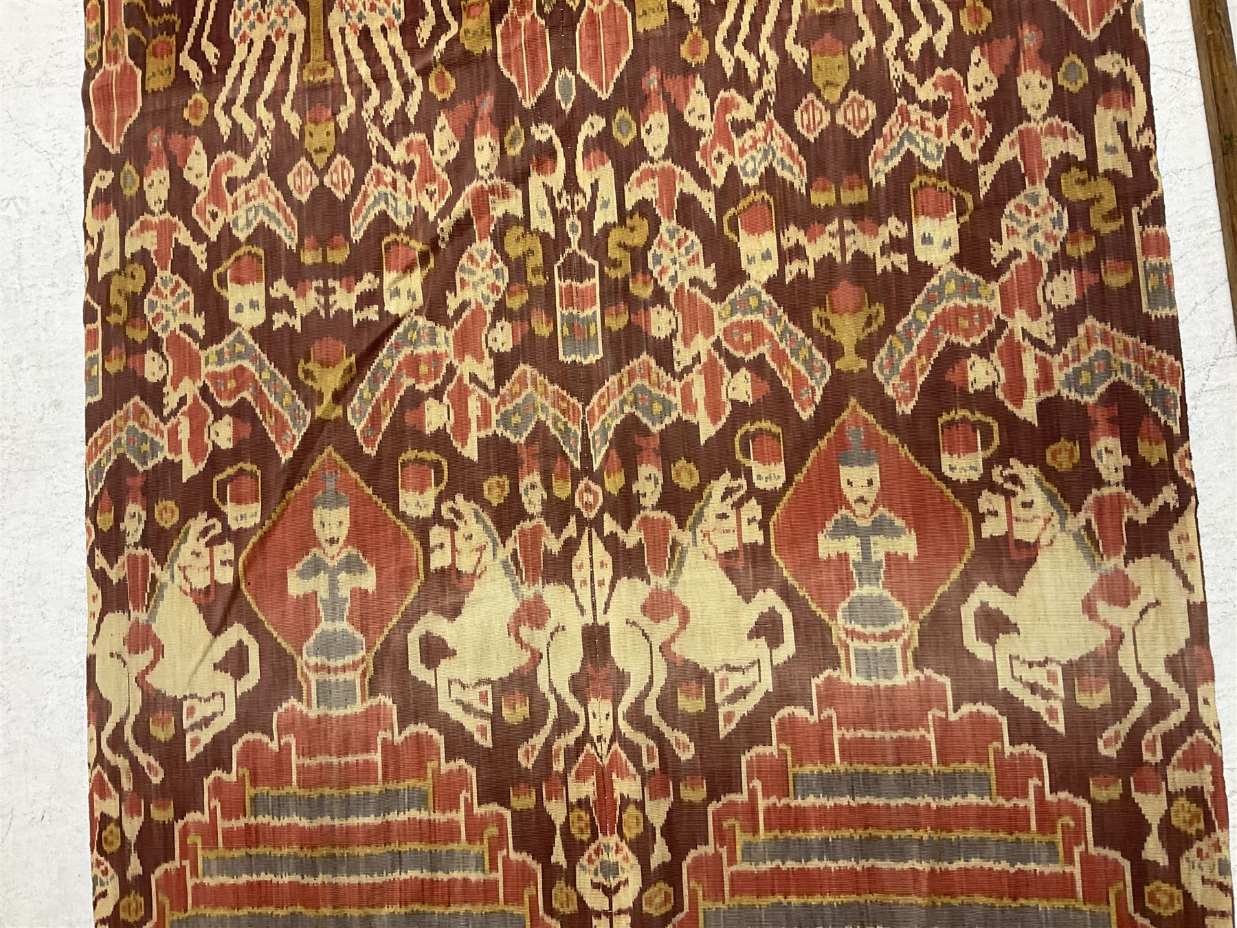 Ikat Man’s Cloth (Hinggi Kombu), East Sumba, Indonesia, woven in red, blue and yellow-brown warp ikat, constructed of two mirroring long panels, the motifs arranged in four bands depicting warrior figures astride rearing horses, high-ranking figures and guardians within geometric borders, on carved wooden wall hanger, L295cm

