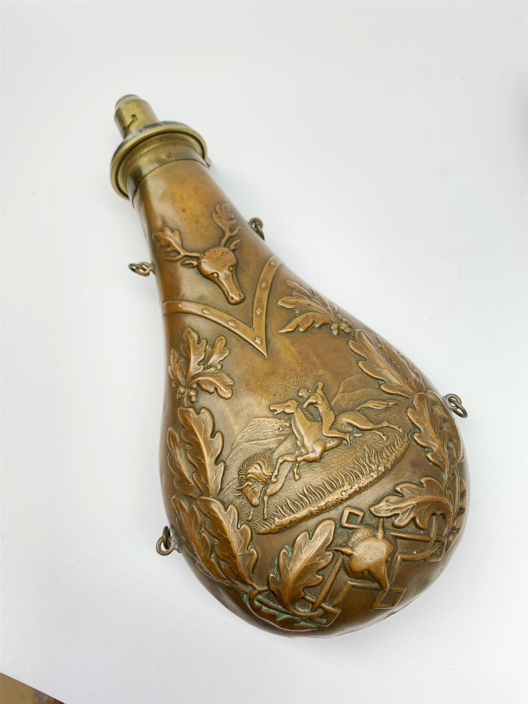 Victorian large copper powder flask embossed with a hunting scene within an oak leaf border with stag and fox heads, the nozzle marked 'Extra Quality Sykes Patent', fitted with four suspension rings H24cm; together with Victorian leather shoulder shot flask with Sykes nozzle L80cm (2)
