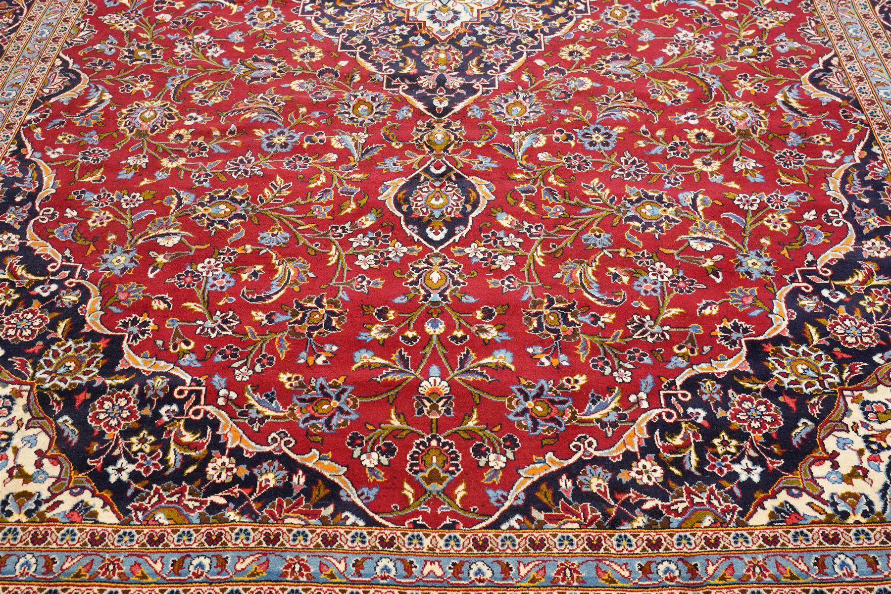 Central Persian Kashan hand-knotted carpet, crimson ground field, with central cusped lozenge medallion, enclosing a palmette and a floral rosette arrangement, surrounding field densely populated with scrolling arabesques, flowering vinery and stylized boteh motifs in tones of indigo, pale blue, ivory and ochre and spandrels of confirming design, flanked by multiple guard stripes, with cotton fringe ends 