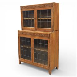 Early 20th-century Arts and Crafts glazed oak bookcase, upper section fitted with a pair of leaded glazed doors enclosing a fixed shelf above a projecting frieze drawer interior with baize-lined writing surface flanked by two compartments, carved with interlaced decoration, lower section fitted with a further pair of leaded glazed doors enclosing a single shelf, raised on short stile feet 