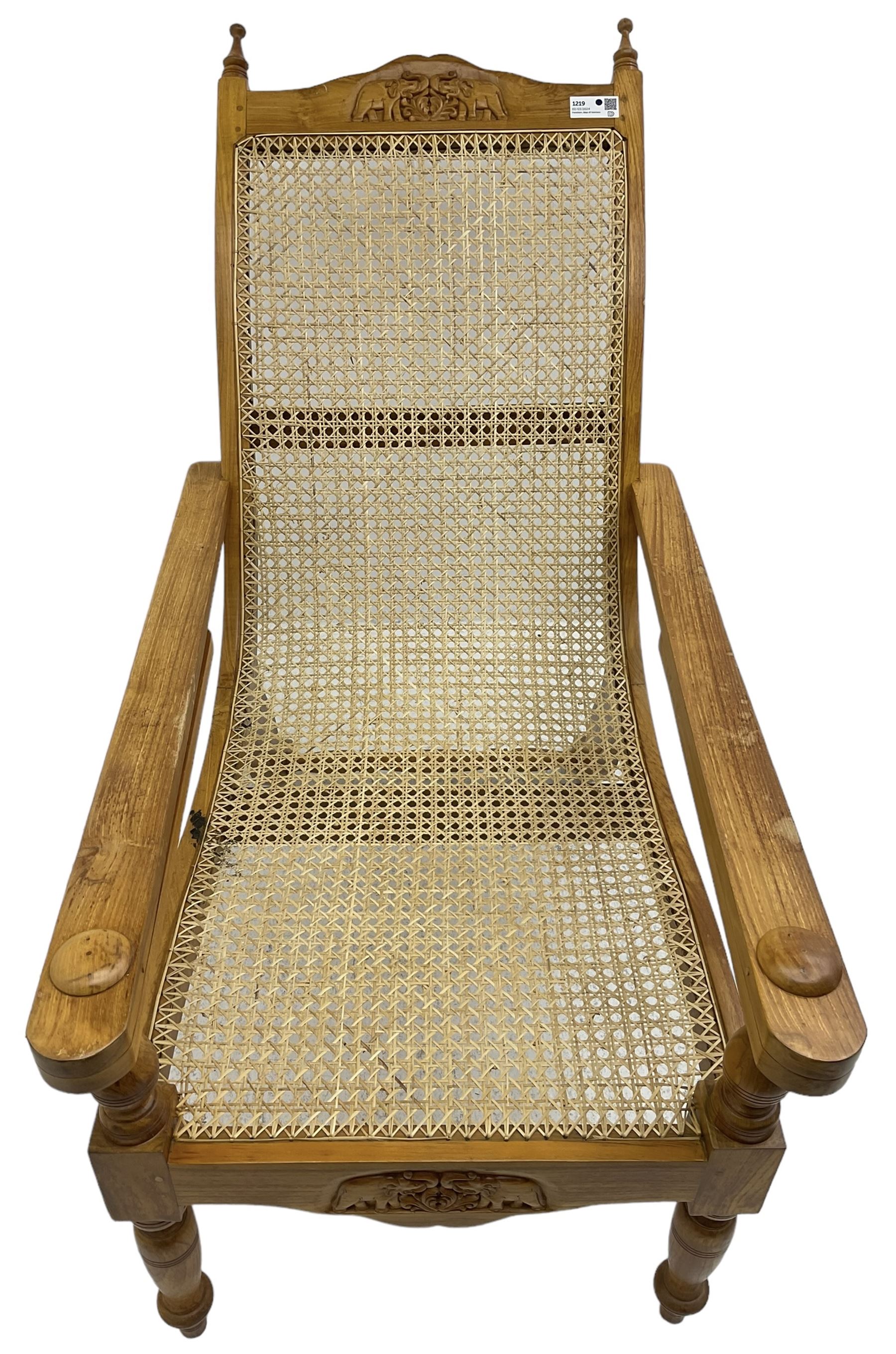Late 20th century teak framed plantation chair, cresting rail and apron carved with two elephants with upraised trunks, flanked by finials, cane back and seat, fitted with hinged extending leg rests, on ring turned supports