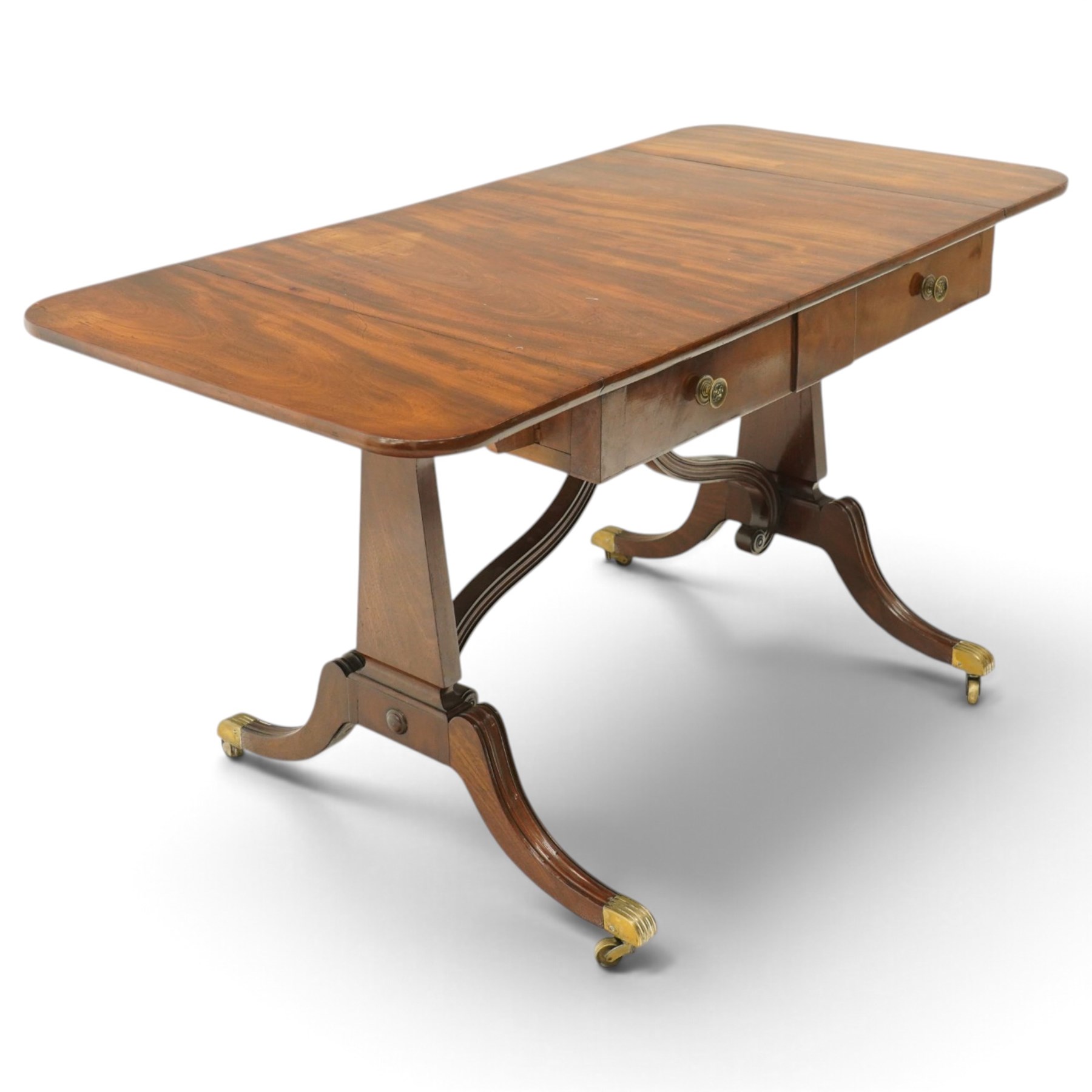 Early 20th century Regency Revival figured mahogany sofa table, rectangular drop-leaf top with rounded corners, fitted with two drawers to each side with brass flowerhead pull handles, raised on twin end supports with splayed reeded legs and brass cups and castors, united by a scrolling and reeded closed stretcher