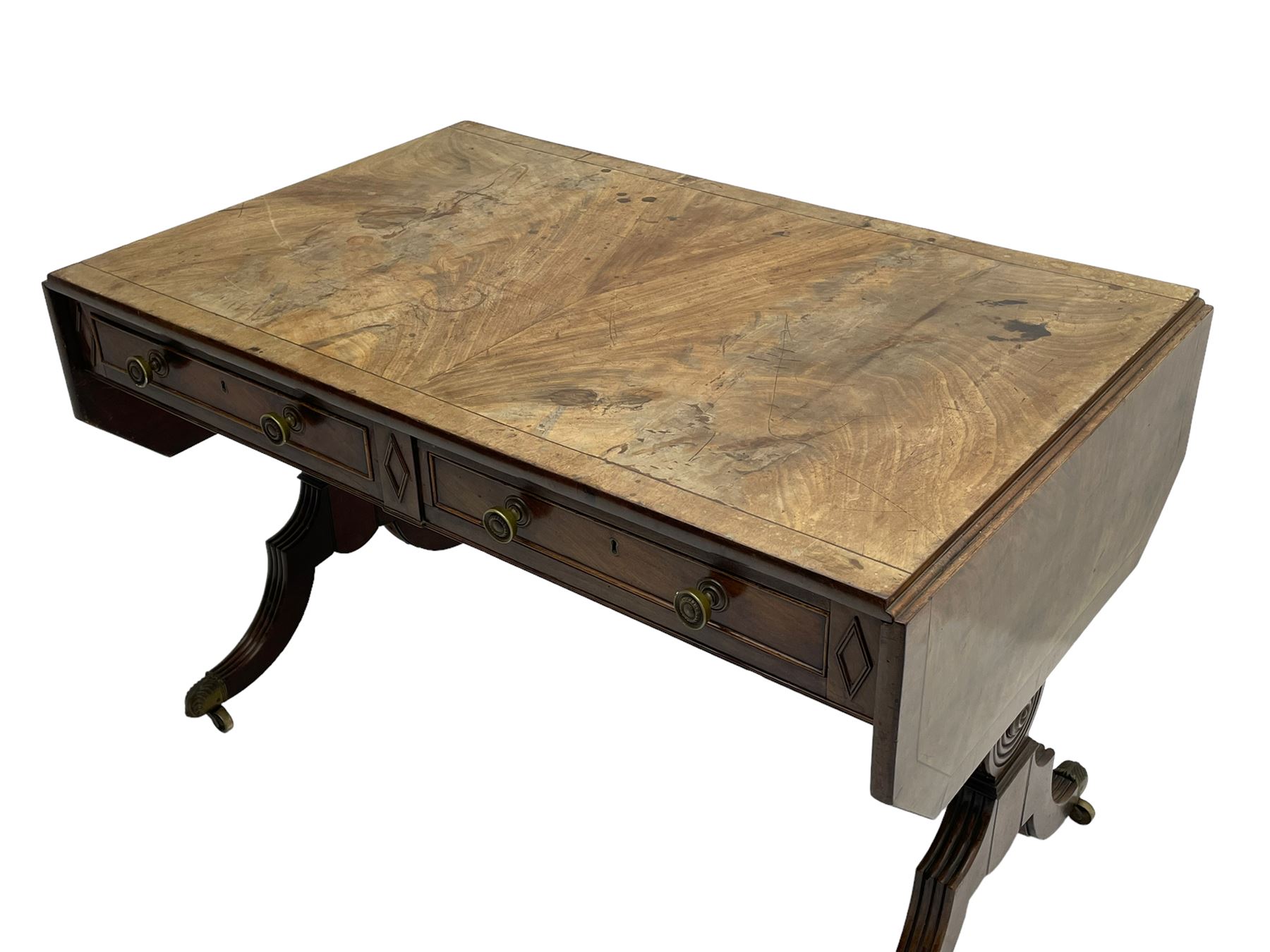 Regency mahogany sofa table, canted rectangular drop leaf top with walnut crossbanding, fitted with two frieze drawers with central lozenge decoration, two opposing false drawers, vasiform end supports with reeded edge and central roundel joined by ring turned and reeded stretcher, splayed supports on scaled brass cups and castors
