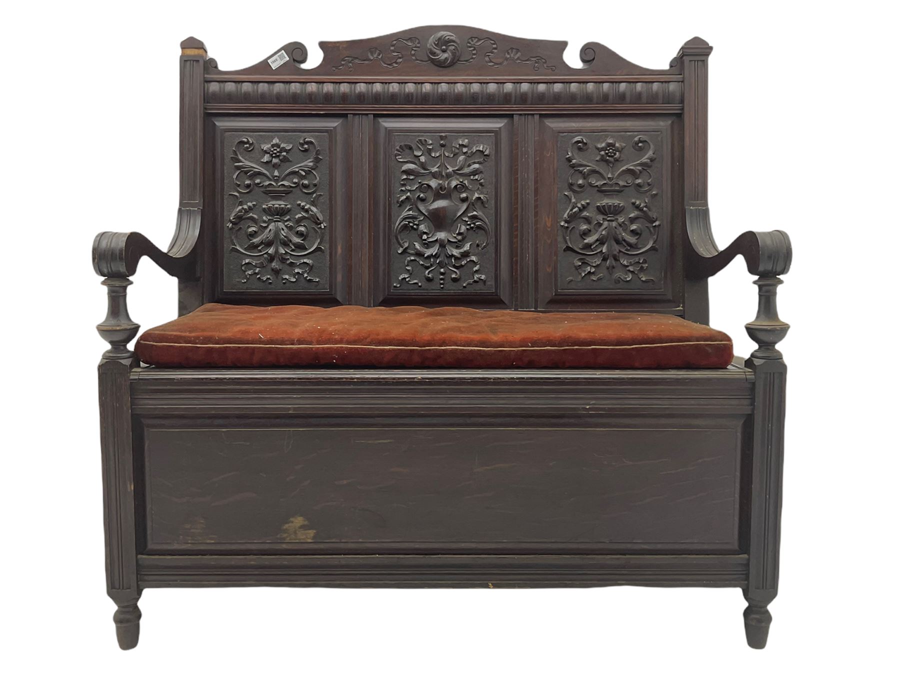 Late 19th century oak settle bench, shaped cresting rail carved with ribbon over triple panelled back, the panels relief carved with scrolled and floral urns and cartouche, hinged box seat, turned front feet, with upholstered squab cushion