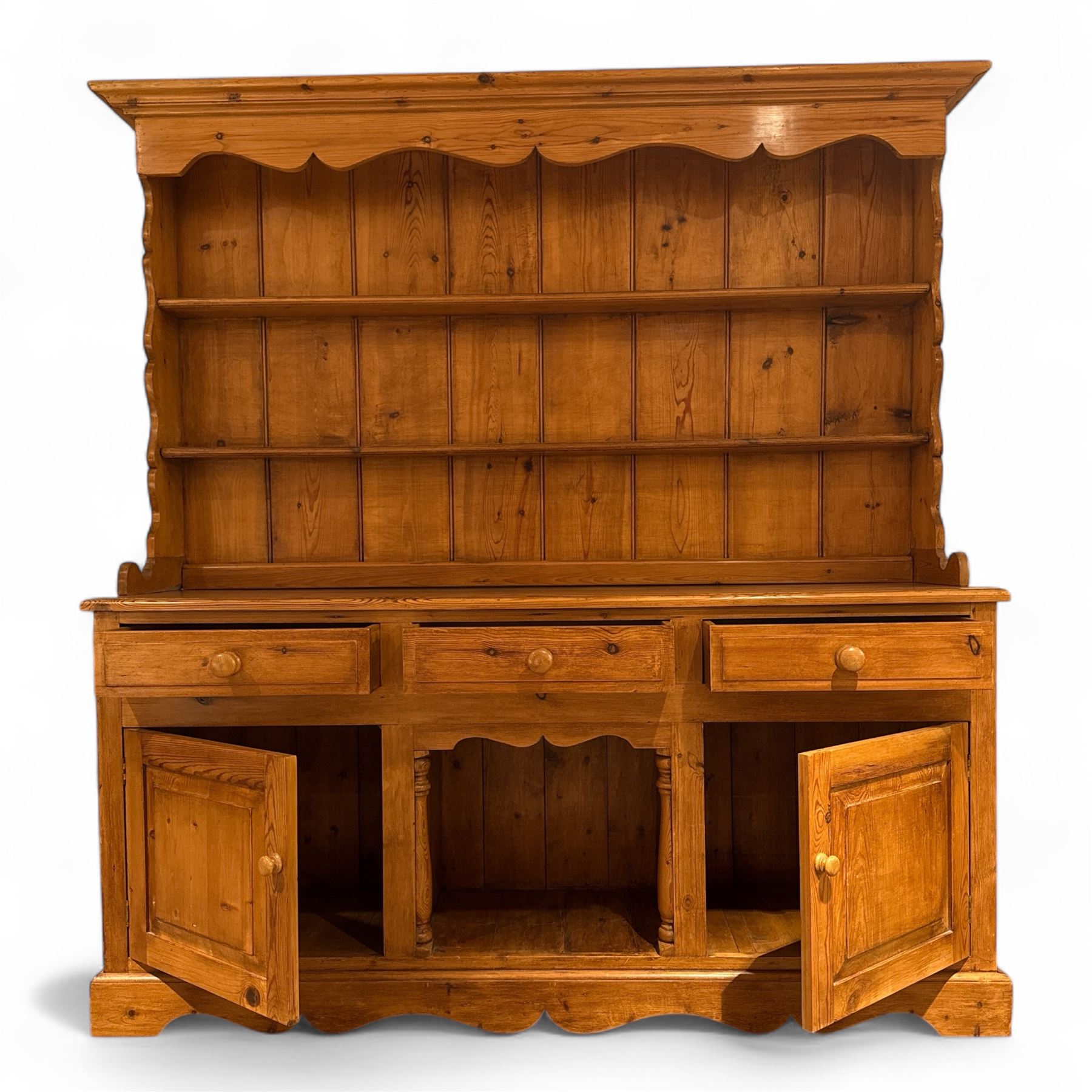 Late 20th century pine dresser, the plate rack with two fixed shelves and shaped apron over panelled back, base fitted with three drawers over two cupboard doors and central open niche flanked by turned uprights, on plinth base with shaped apron