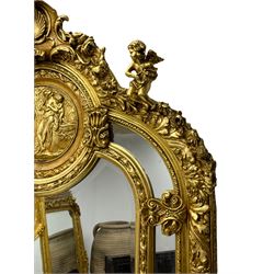 Large Italian Baroque design gilt wall mirror, the shell pediment over circular panel depicting classical female flute player with dancing putto within a tree landscape, the stepped arched upper frame surmounted by musical instrument playing putto and trailing flower heads, central bevelled mirror plate surrounded by plain segmented mirror plates, acanthus leaf moulded outer frame and foliate moulded inner slip